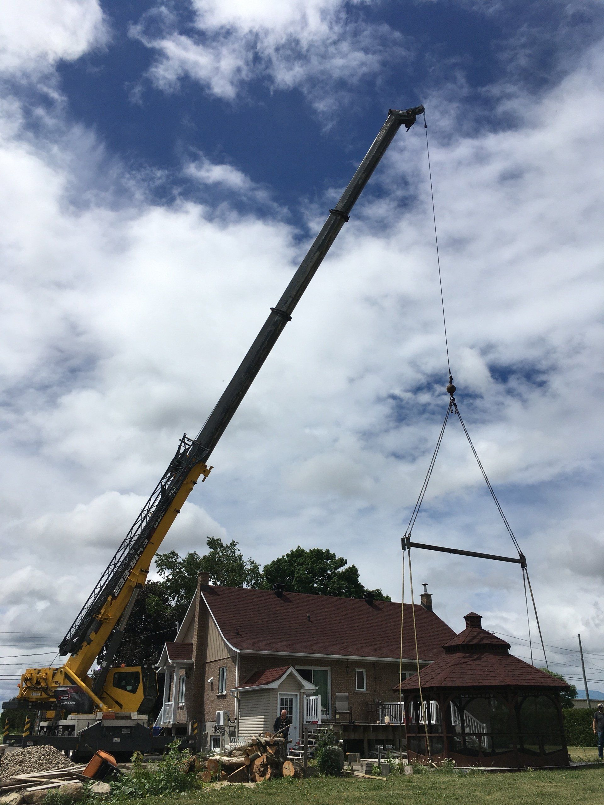 Grue soulevant une structure métallique près d'une maison et d'un belvédère sous un ciel nuageux.
