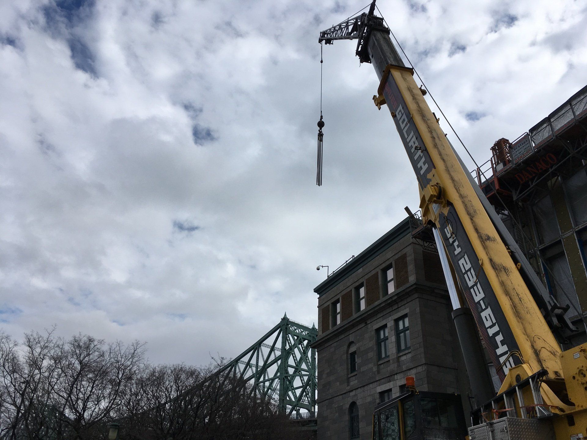 Une grue soulevant un long objet près d'un bâtiment en pierre et d'un pont vert contre un ciel nuageux.