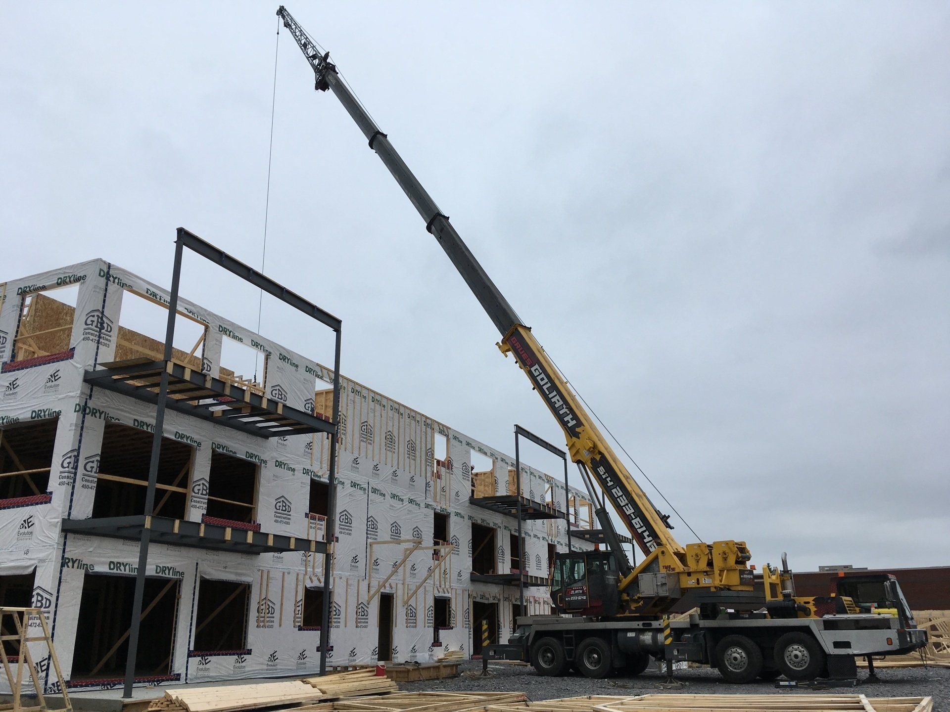 Une grue soulève une poutre métallique sur un chantier de construction à plusieurs étages ; ciel nuageux au-dessus.