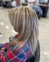 Woman with blonde highlights in a hair salon, wearing a red and blue plaid shirt, sitting in a chair.