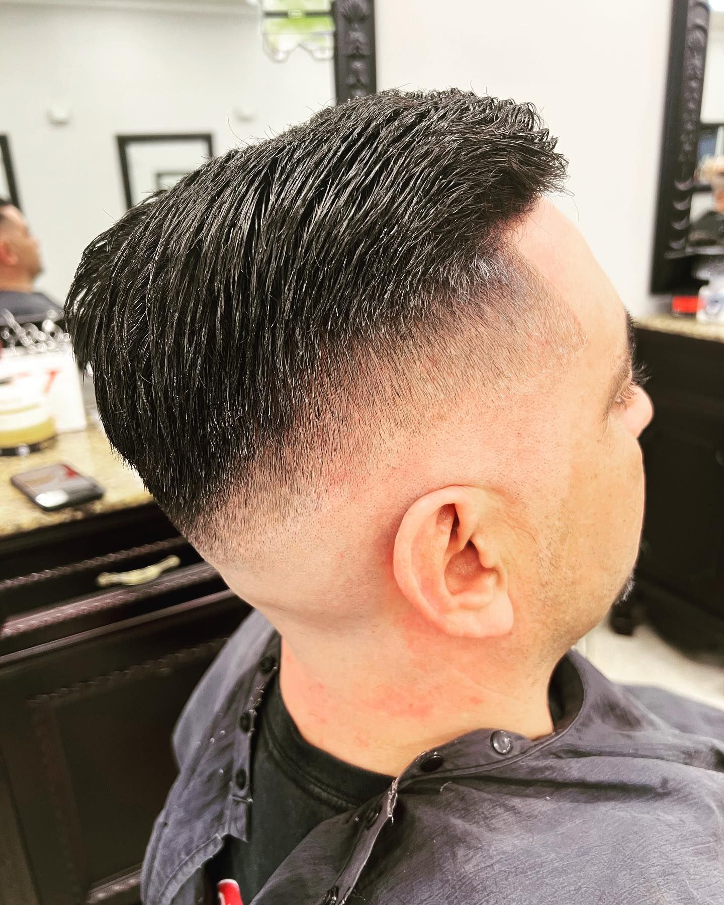 Man with dark hair, faded sides, at a barber shop. He is wearing a black cape.