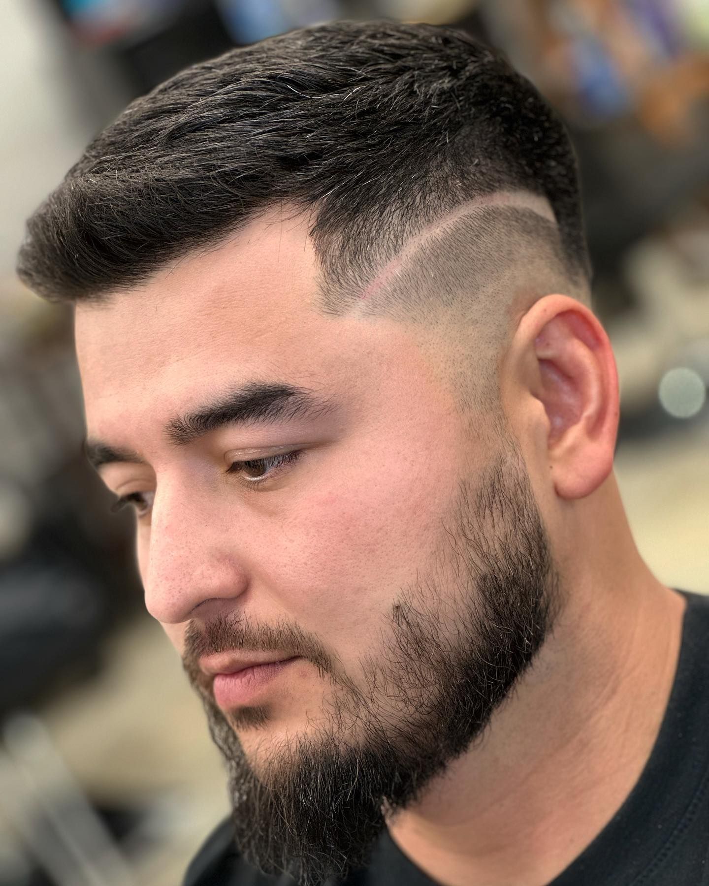 Man with dark hair, faded sides, and beard, looking down; indoors.