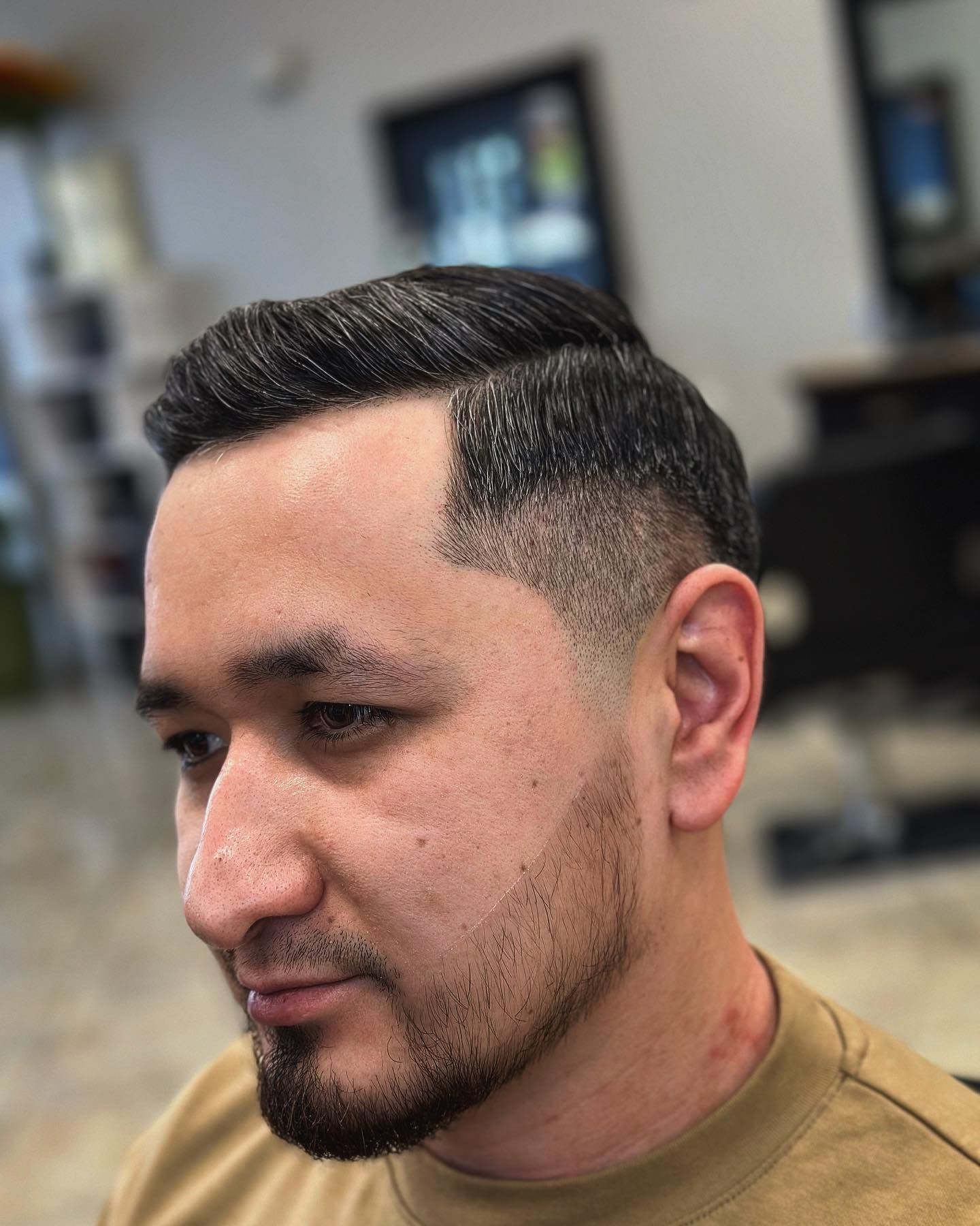 Man with a dark, slicked-back haircut and beard, looking to the side. Faded sides. Indoors.