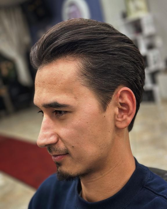 Man with dark hair styled back, faded sides, and stubble. Indoors, profile view.