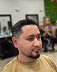 Man with dark hair, fade haircut, beard, smiling in a barbershop.