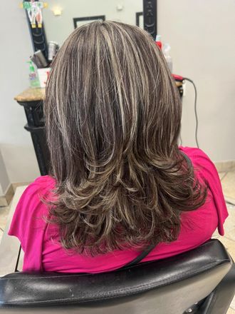 Woman in pink top with layered brown hair with blonde highlights, in a salon.