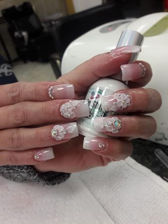 Hands with square acrylic nails, ombre white to pink, 3D flower and rhinestone accents. Holding a nail product bottle.