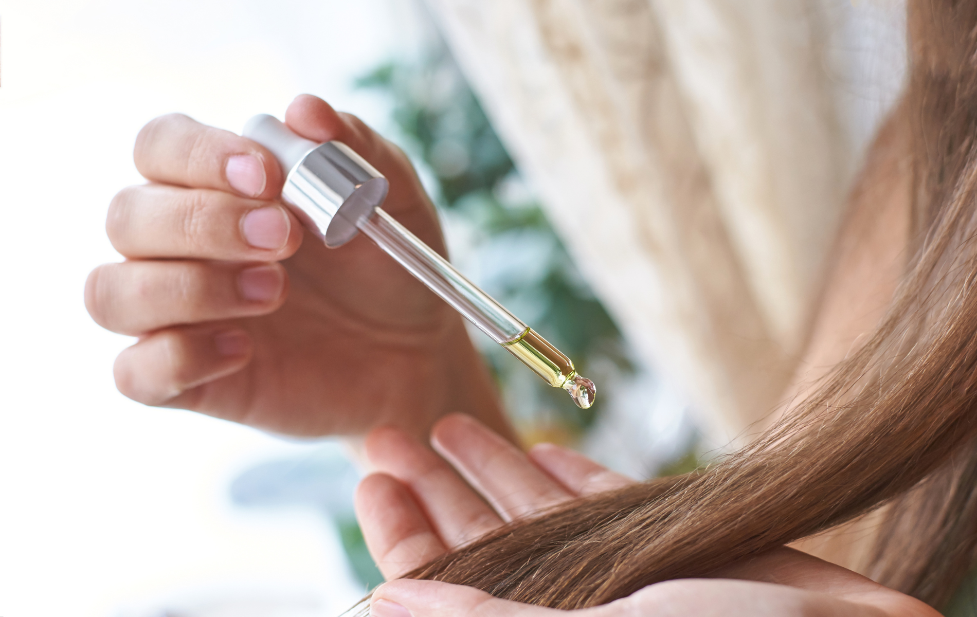 Person applying oil from a dropper to strands of brown hair.