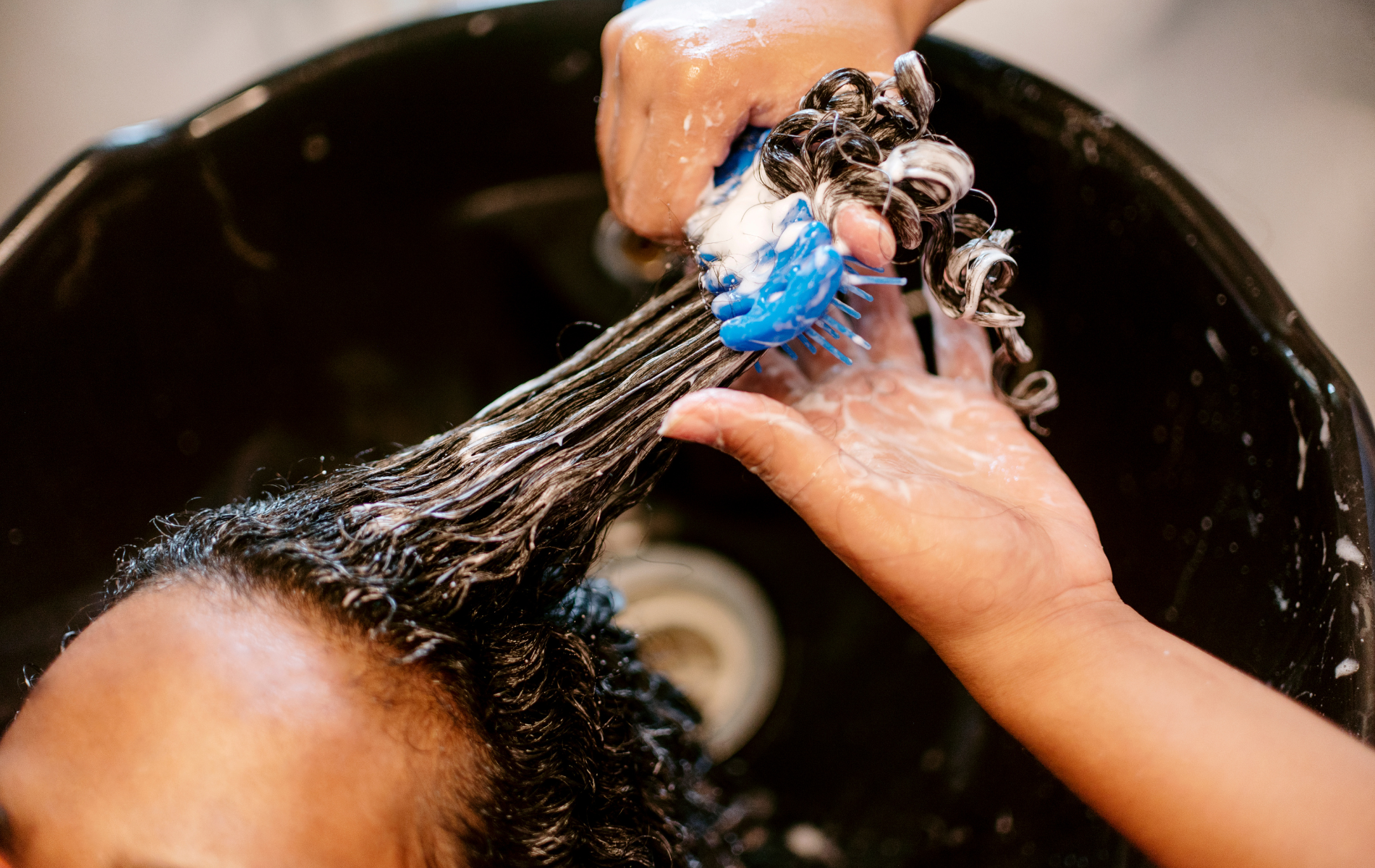 Person having hair washed in a salon sink; a stylist uses a blue brush and suds.