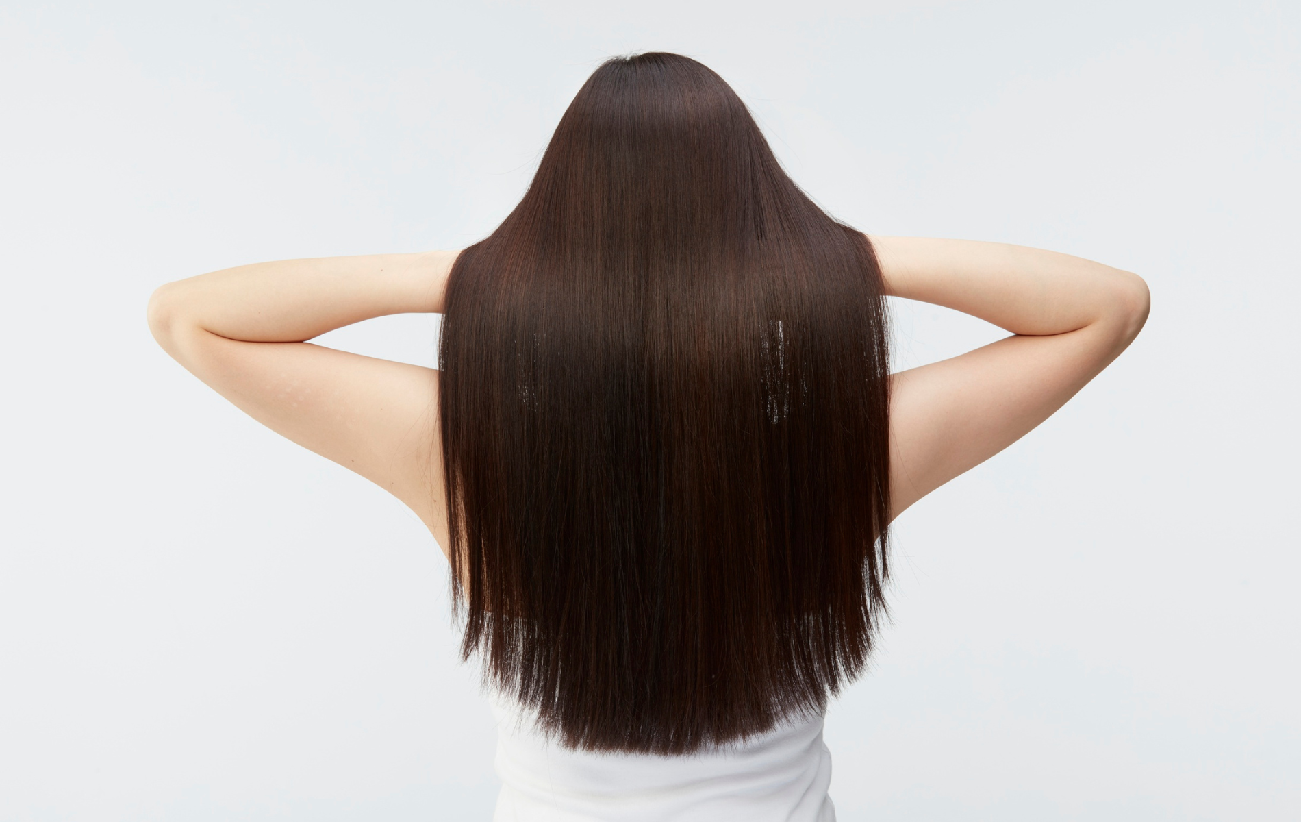 Woman with long, straight, dark brown hair, arms raised to head, standing in front of a white background.