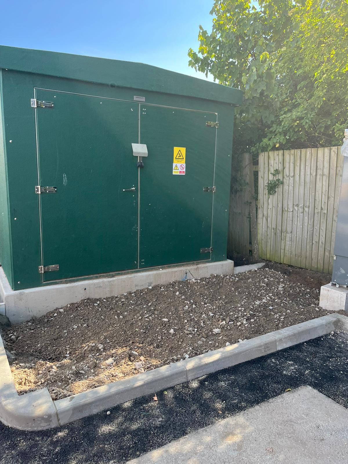A green building with a yellow warning sign on the door is sitting next to a fence.
