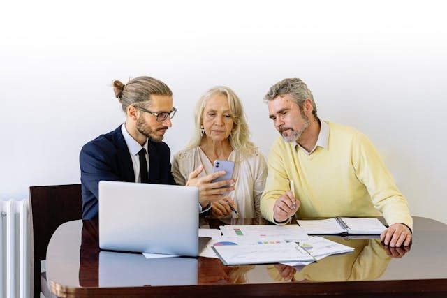 a property manager shows two people something on a phone while sitting in front of a laptop with other documents and reports next to it