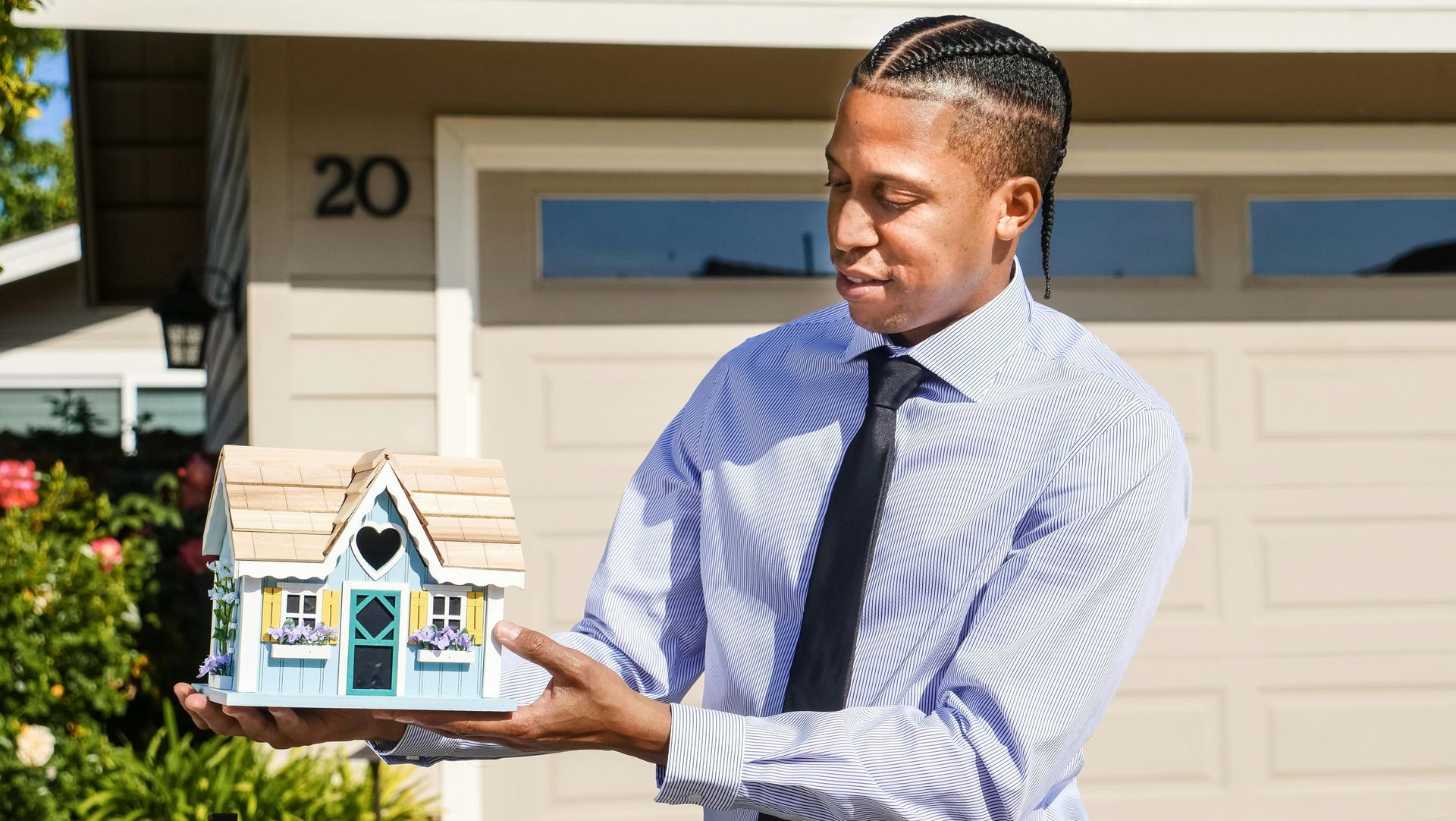 a man in a dress shirt and tie holding up a model house in front of a garage