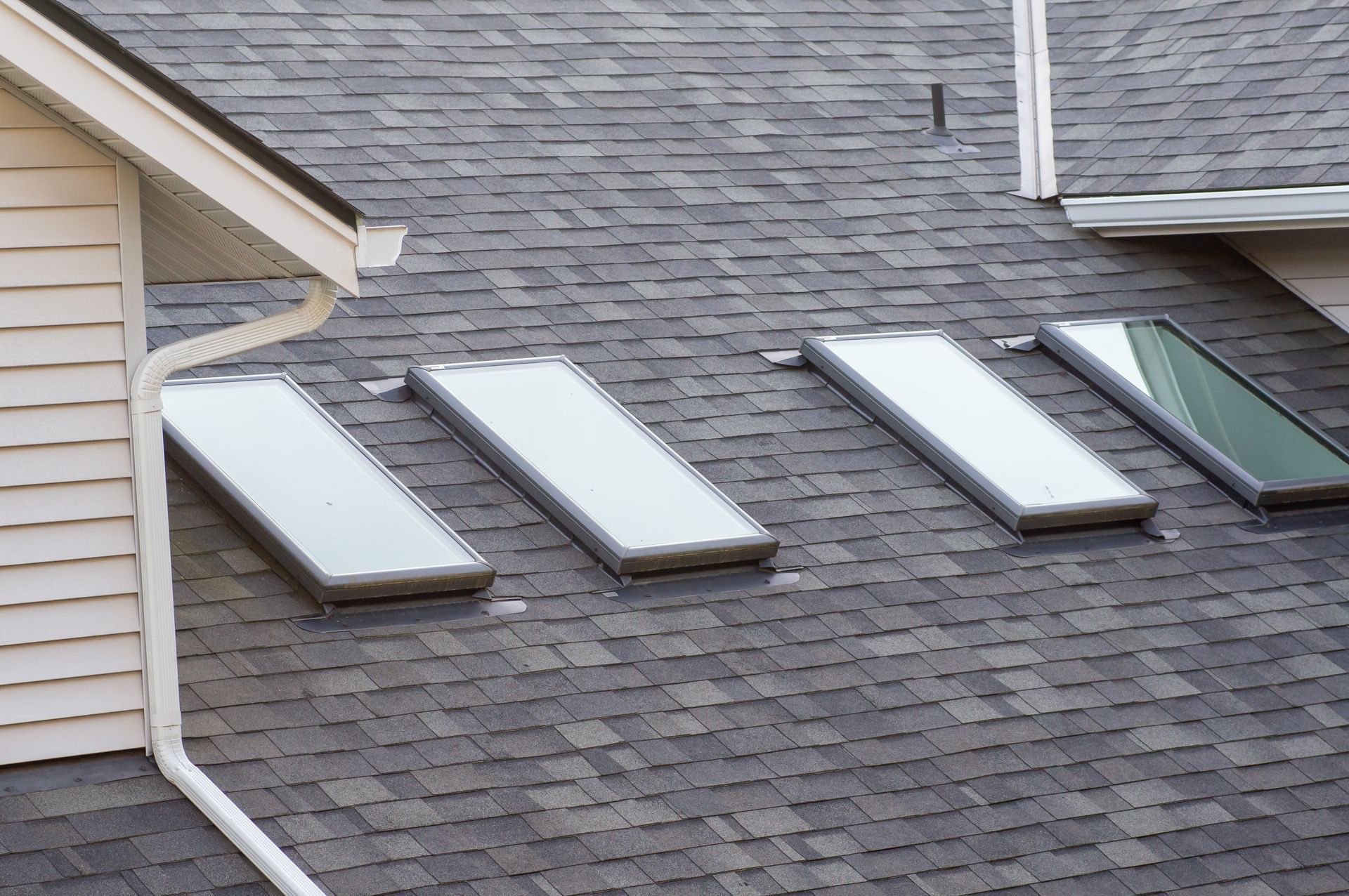 Roof with four skylights on gray shingles beside a beige house exterior