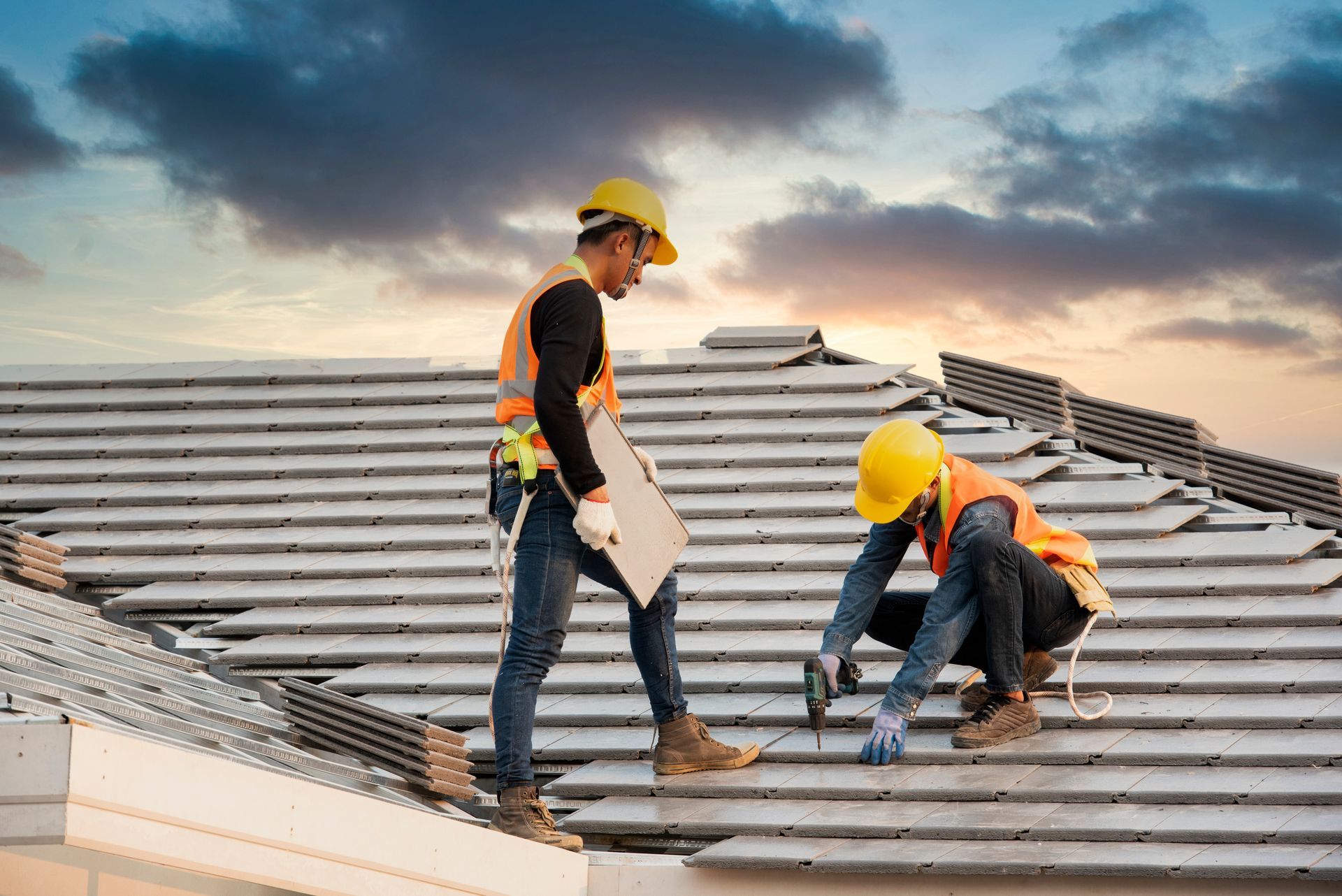 Two construction workers installing metal roofing on a sloped roof at sunset