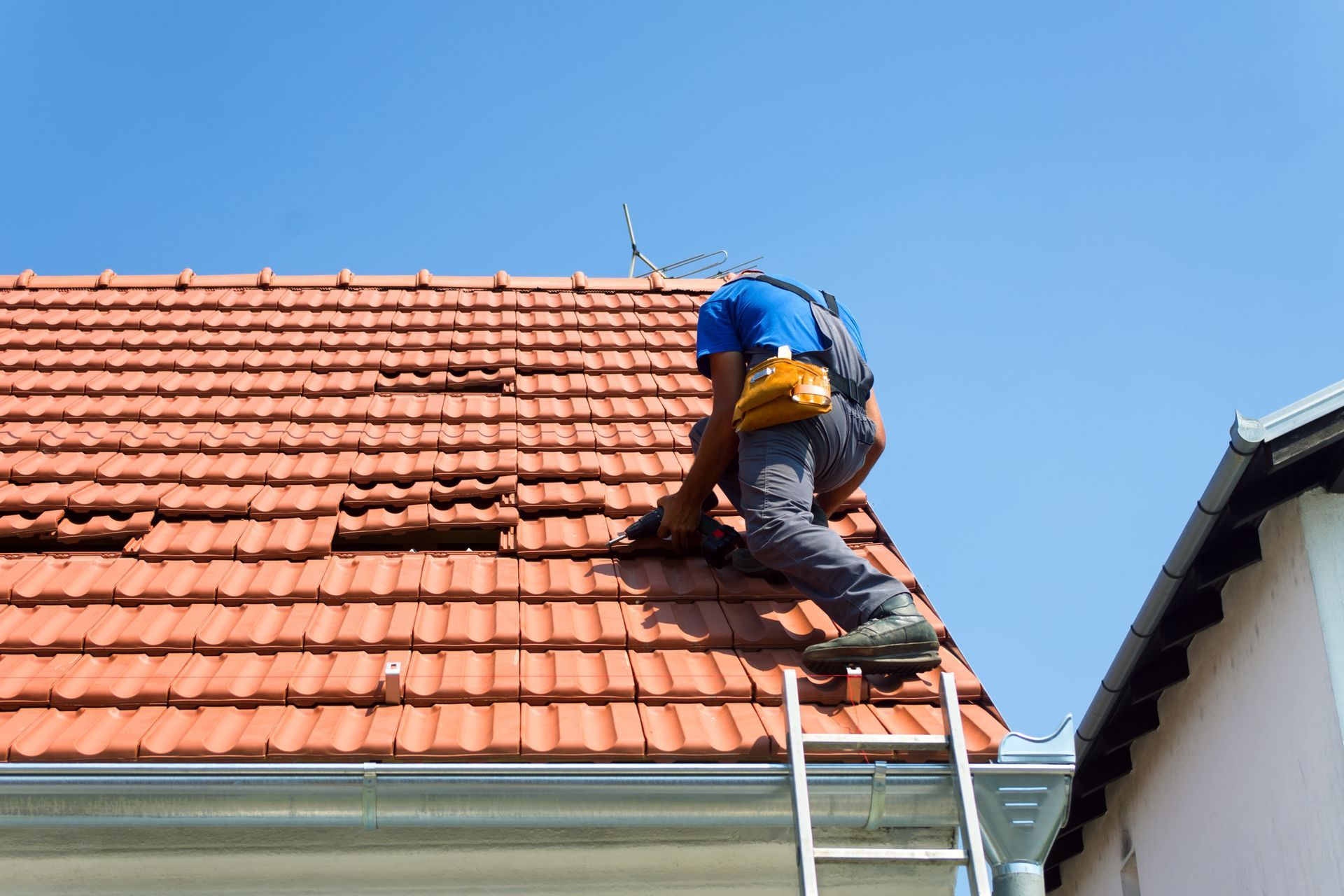 Worker on a ladder repairing red roof tiles under a blue sky