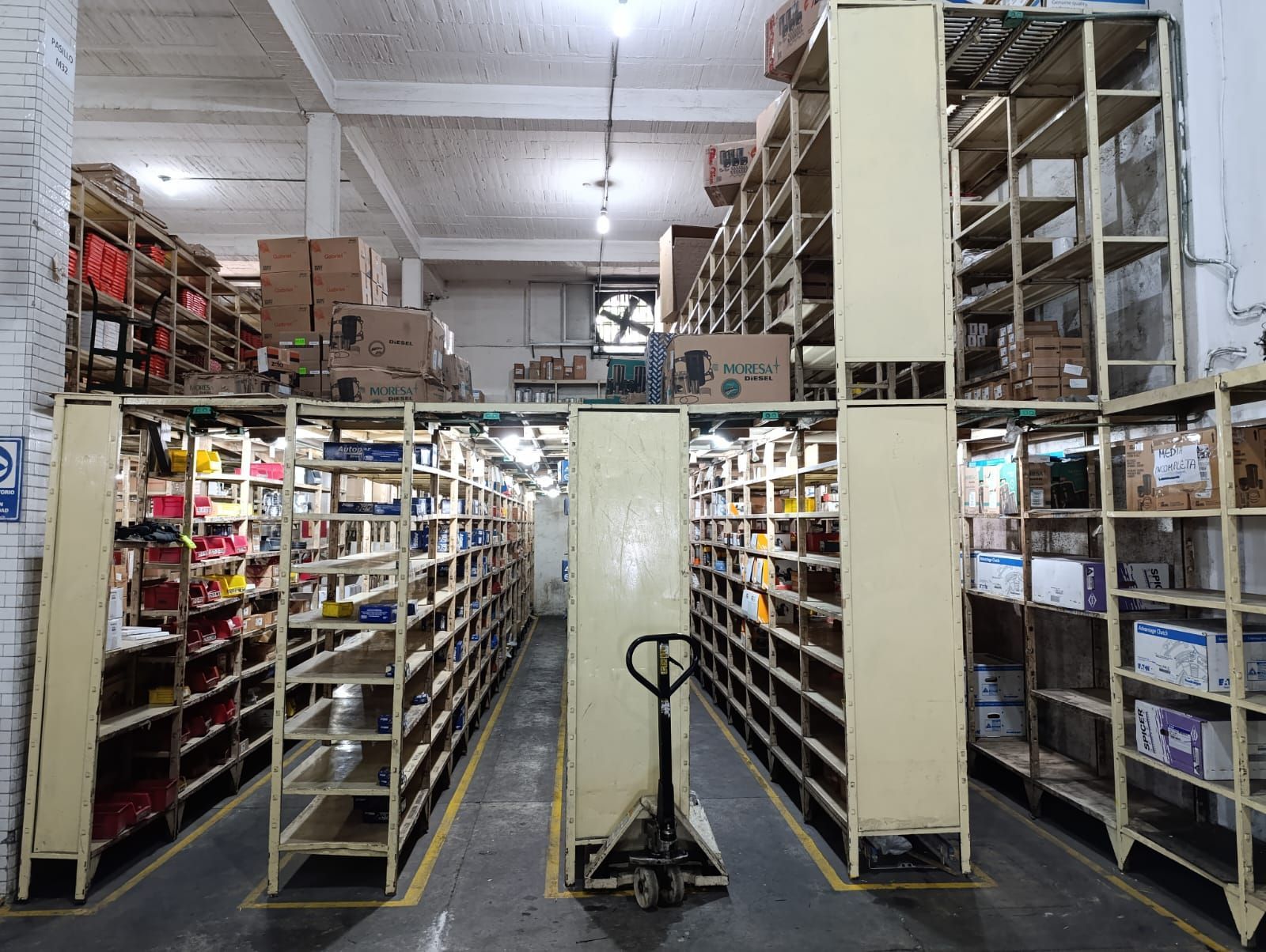 Interior de almacén con estanterías metálicas repletas de cajas y suministros. Carretilla de mano en el pasillo.