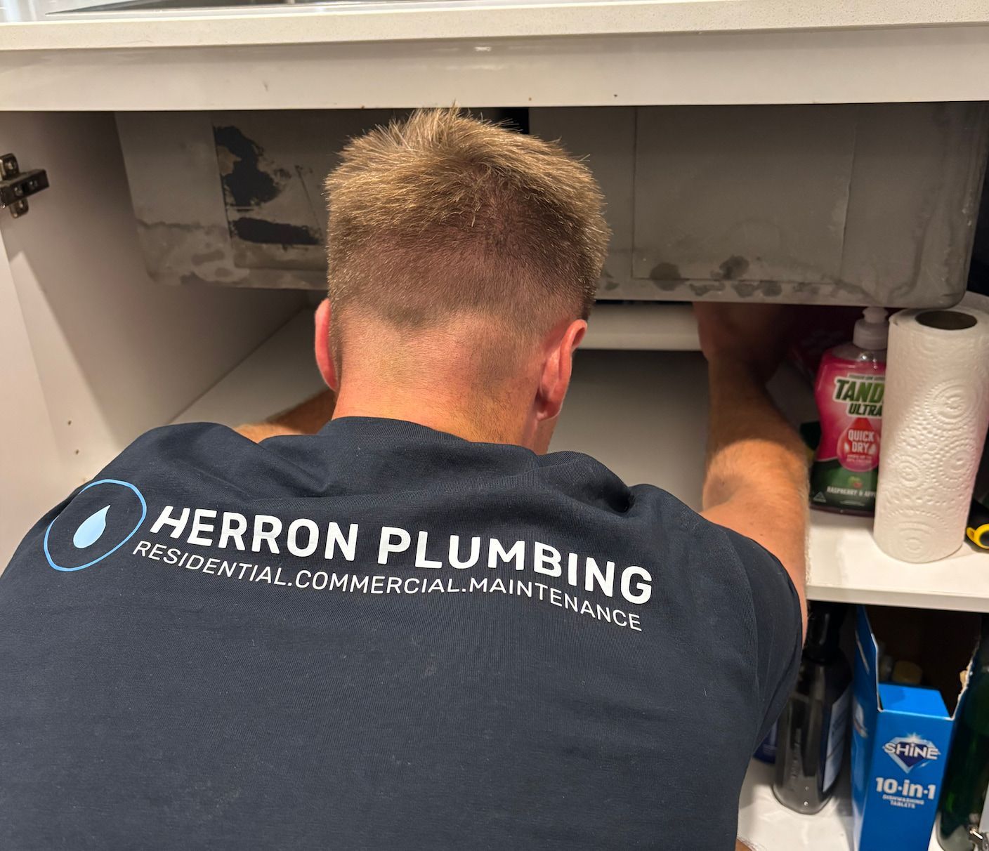 A Person is Cleaning a Drain Under A Sink — Herron Plumbing in Flinders, NSW