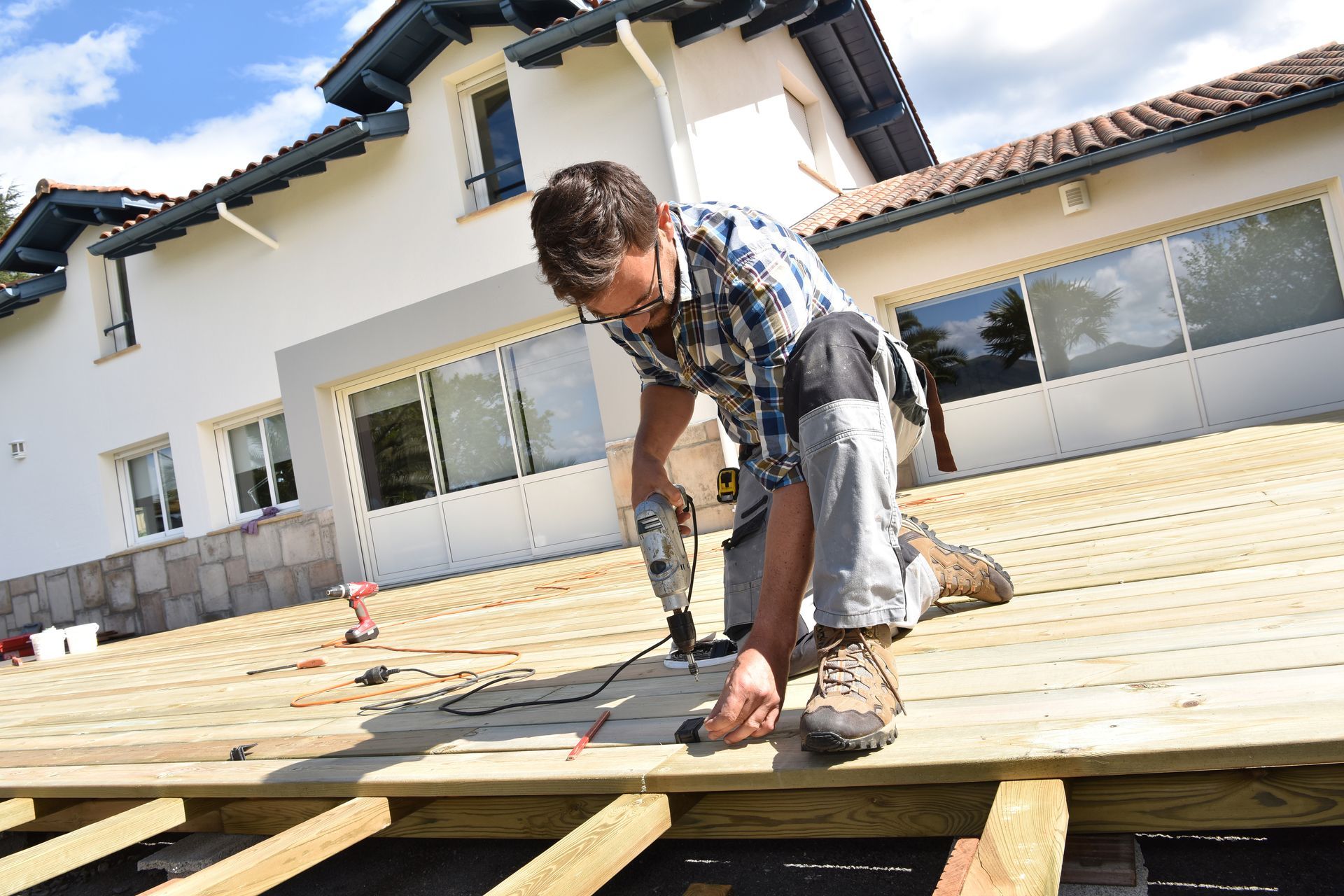 Man building a wooden deck outdoors, using a drill. Light wood, white house in background.