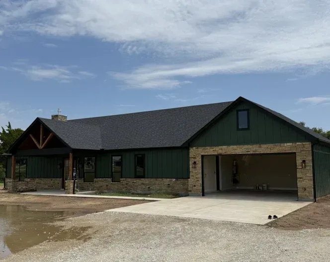 Green-sided house with attached garage, tan brick facade, dark roof, cloudy sky.