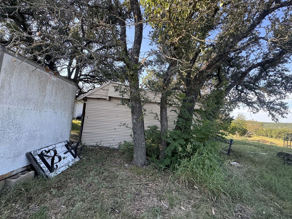 A weathered shack nestled among trees, with a fallen sign on the ground. Overgrown grass and a hilly background.