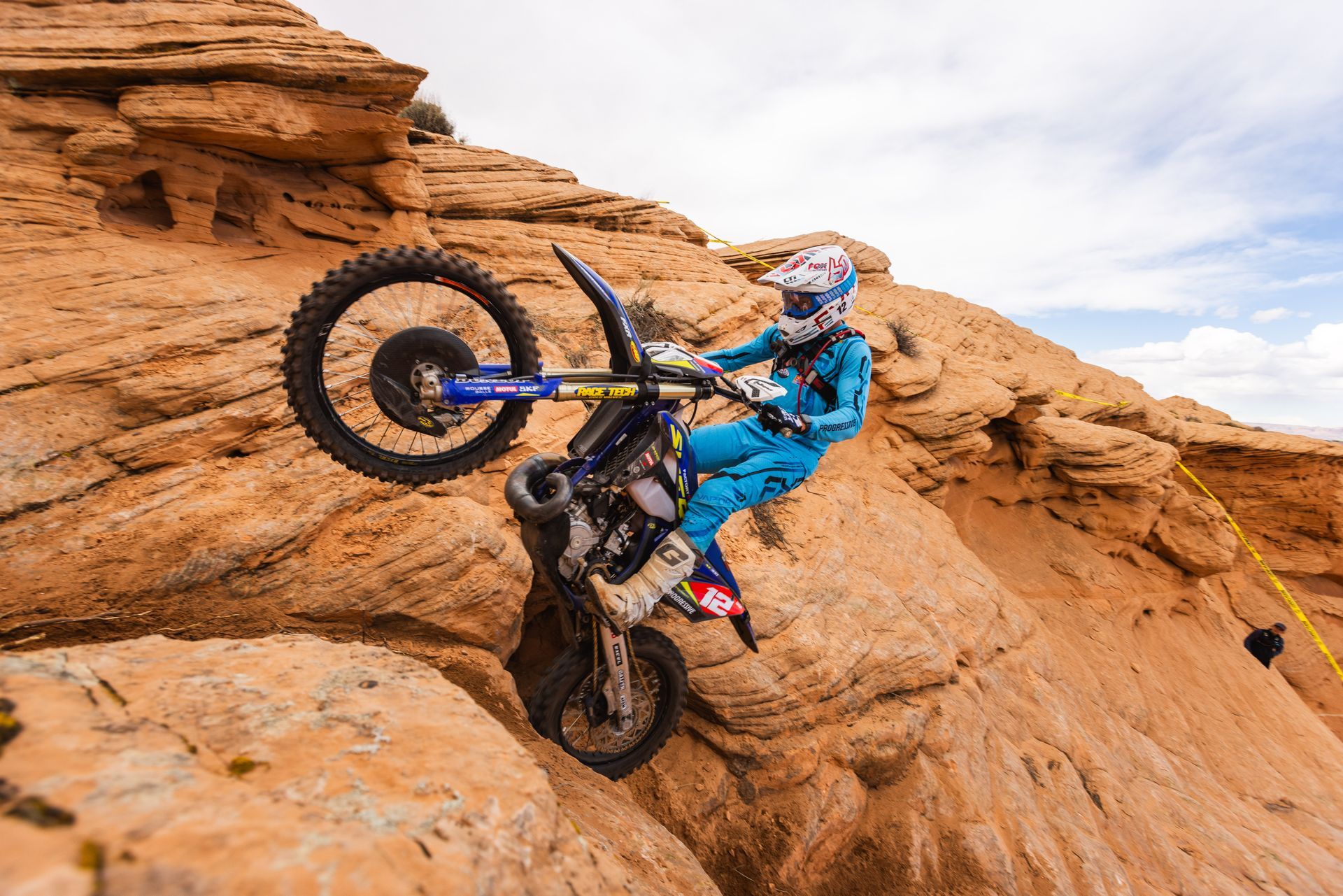 Motorcycle rider on a blue Yamaha, navigating a steep, rocky sandstone slope, wheelie up.