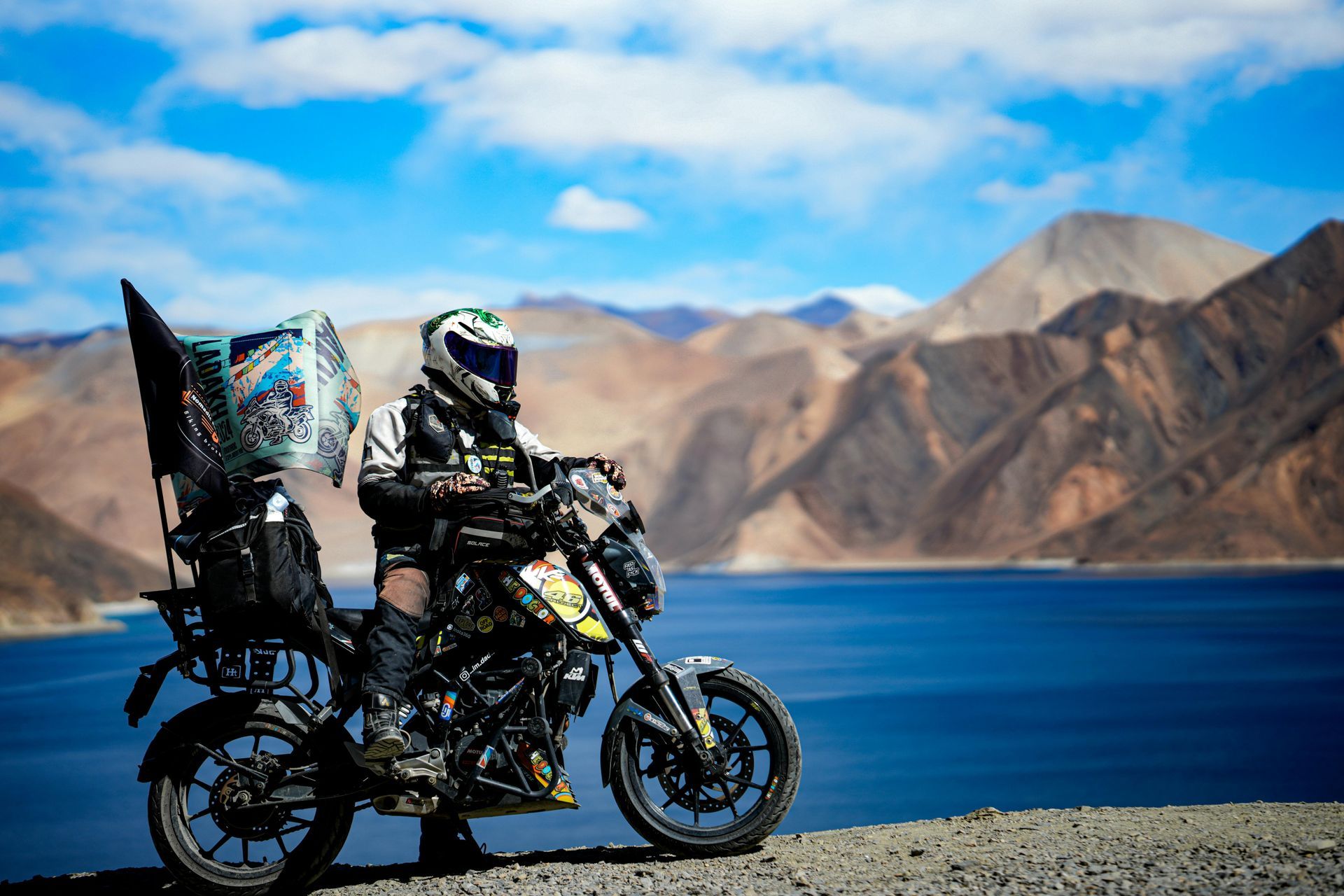 A motorcyclist in gear sits on a bike by a deep blue lake, set against a backdrop of rugged, tan mountains.