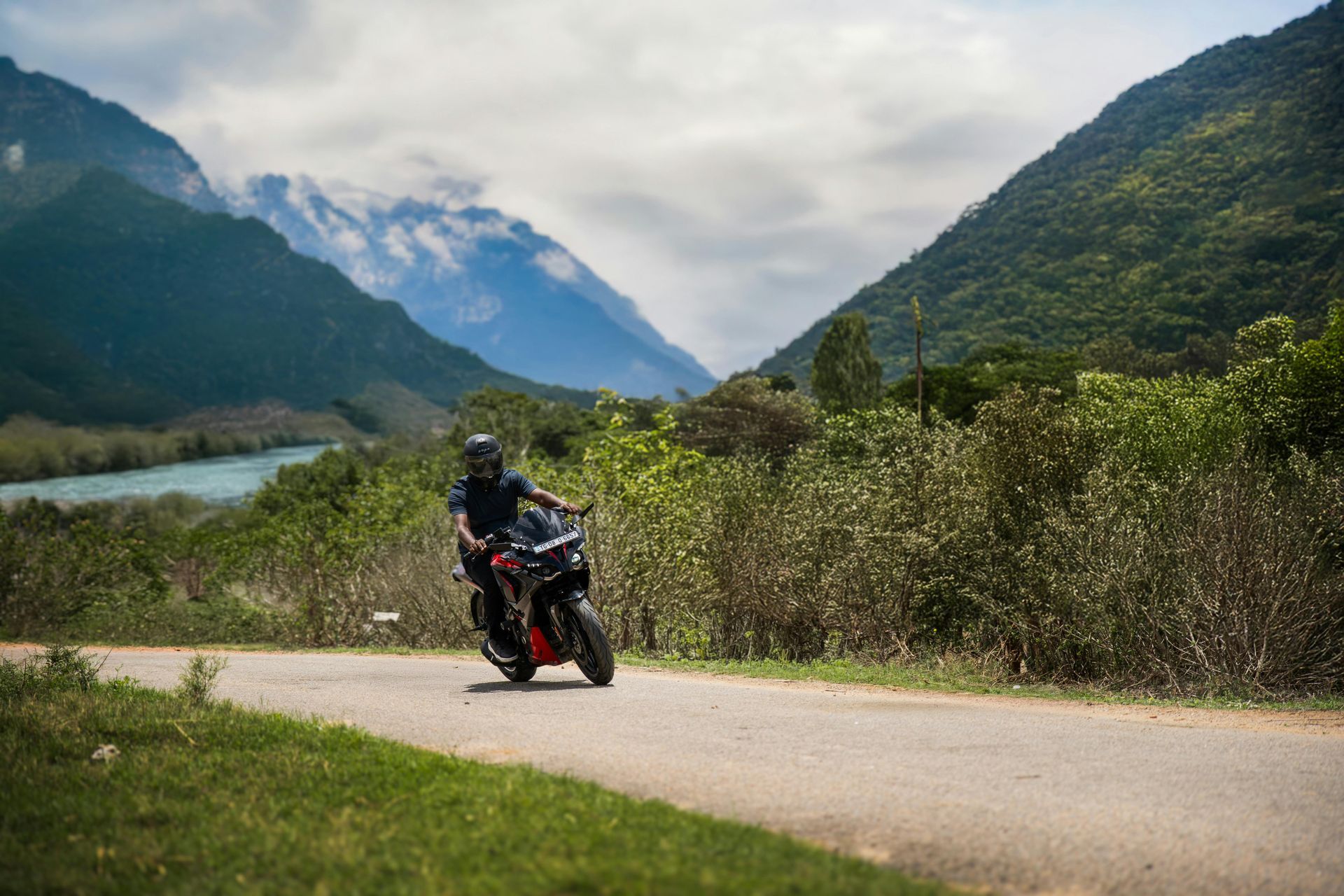 Motorcyclist rides a black and red sport bike on a mountain road with a river and mountains in the background.