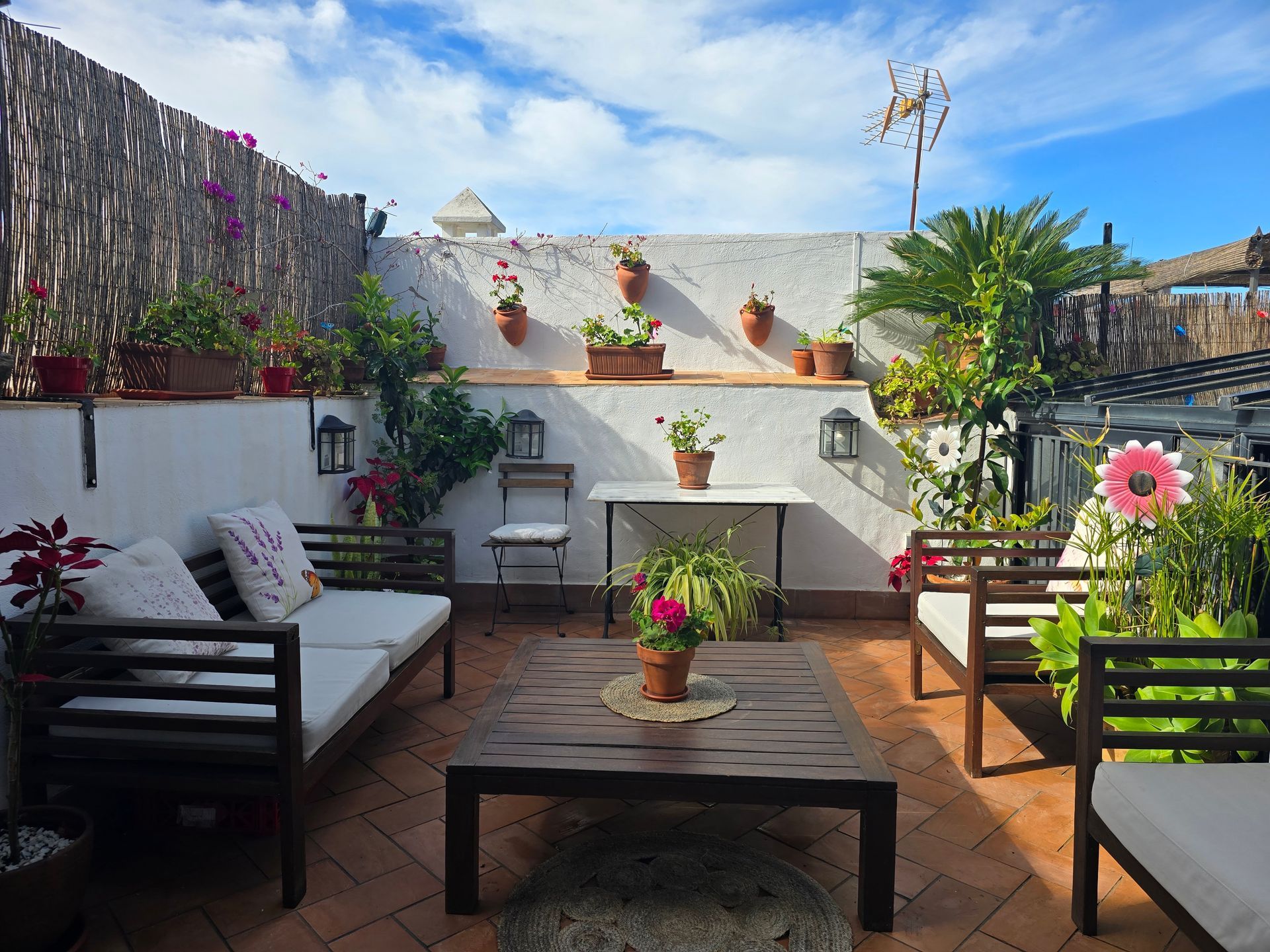 Patio en la azotea con asientos, plantas y una mesa pequeña. Luz tenue al atardecer y cielo azul con nubes.