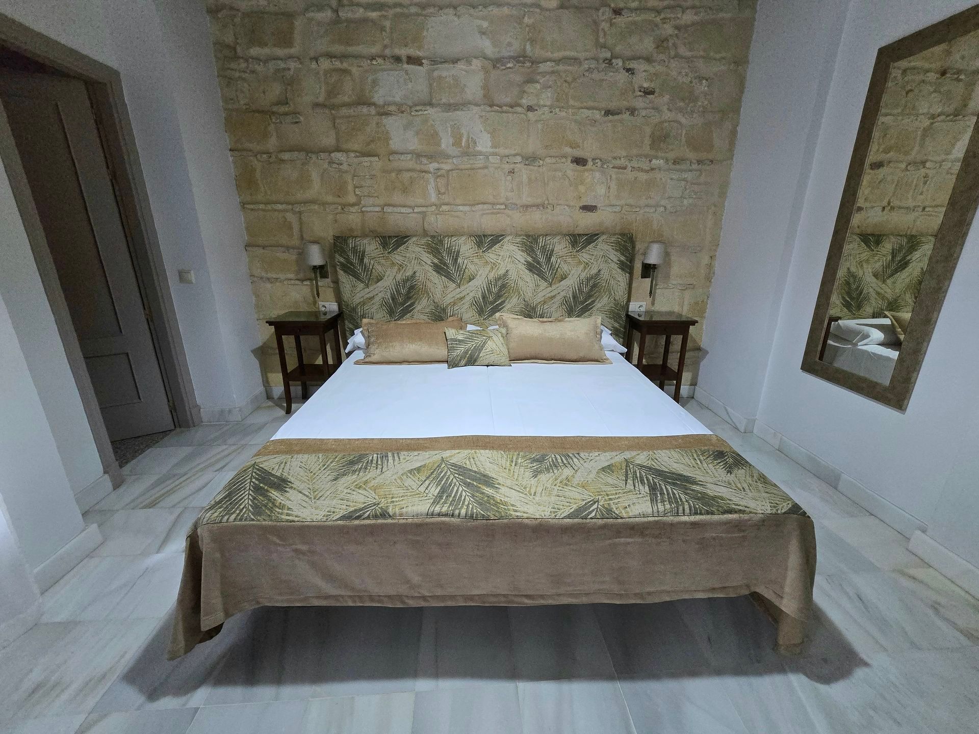 Dormitorio con pared de piedra, cama con funda y cabecero estampado, mesitas de noche, espejo.