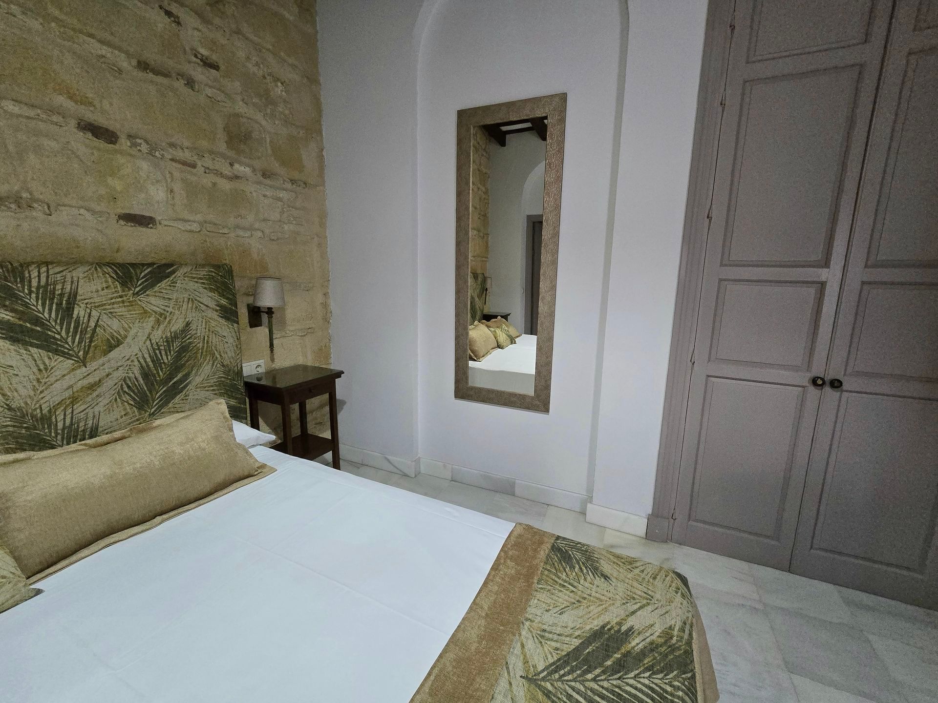 Dormitorio con pared de piedra, cama con cabecero estampado y espejo alto. Tonos claros, texturas suaves.