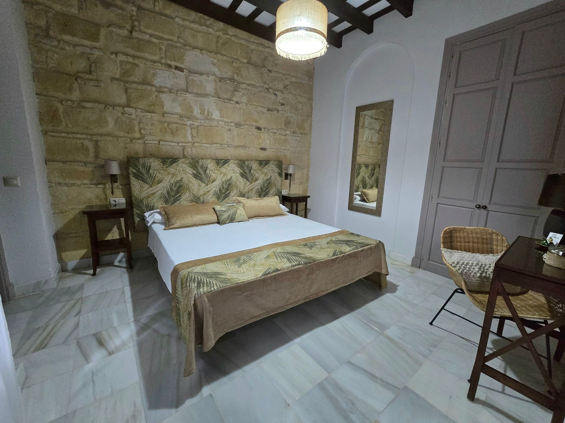 Dormitorio con pared de piedra, cama con cabecero y manta estampada, suelo de madera, mesas auxiliares