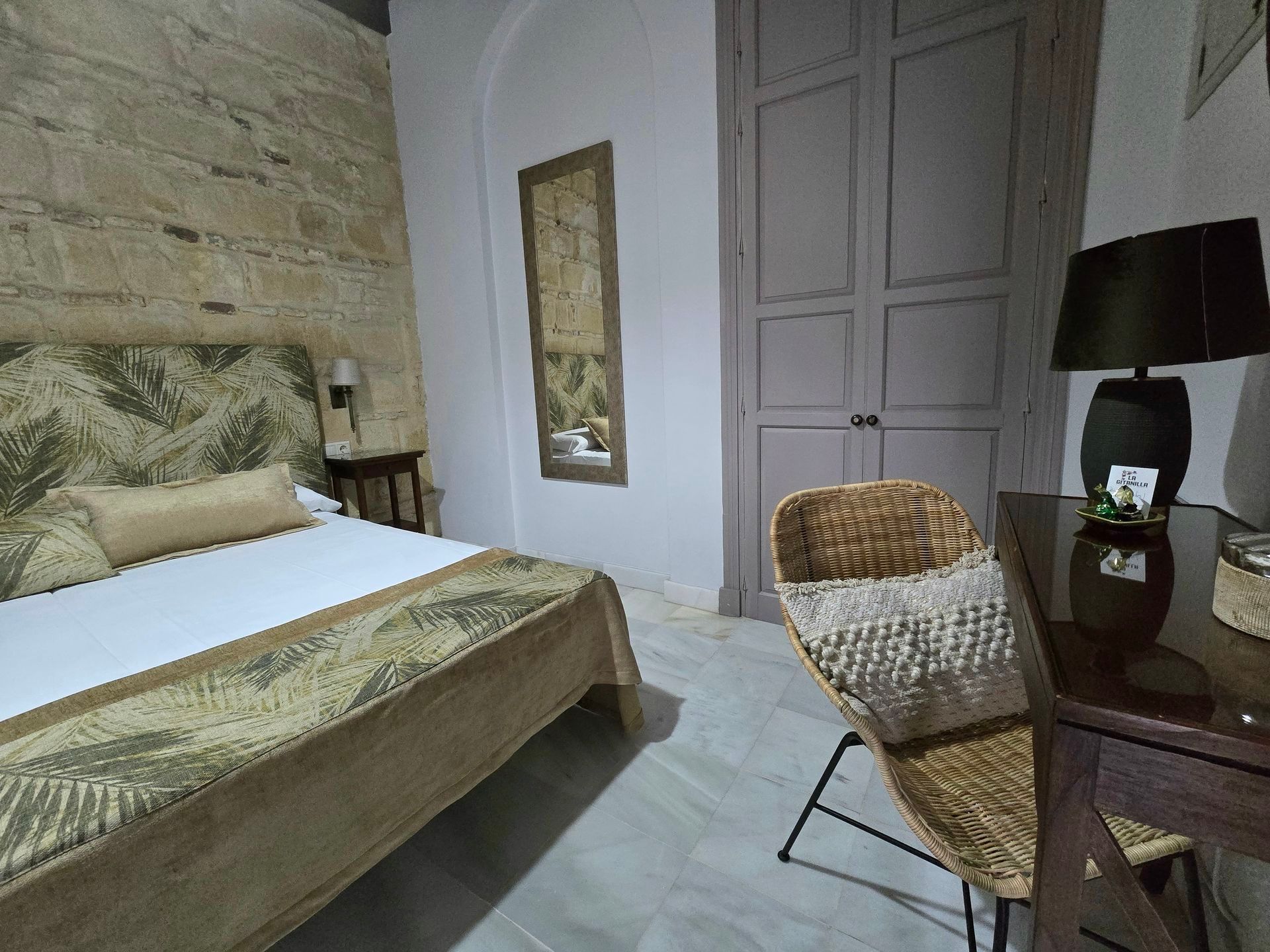 Dormitorio con pared de piedra, cama con funda estampada, escritorio, silla de mimbre y armario.