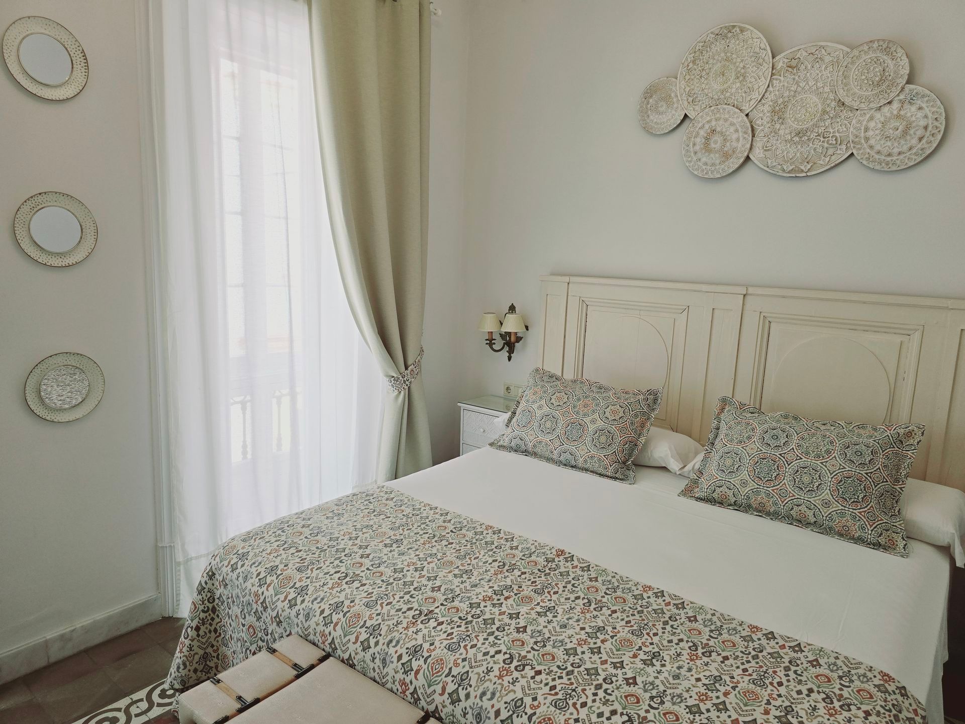 Dormitorio con cama blanca, ropa de cama estampada y decoración en las paredes. Ventana con cortinas a la izquierda.