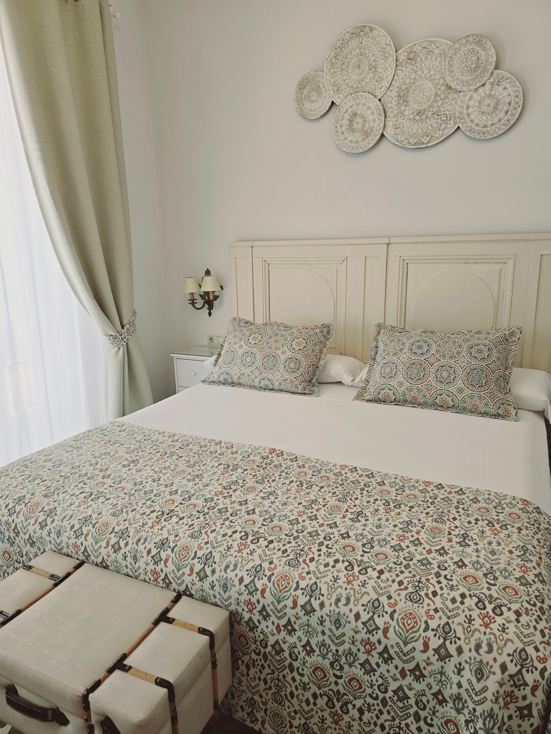 Dormitorio con cama blanca, ropa de cama floral, arte decorativo de nubes y cortinas beige.