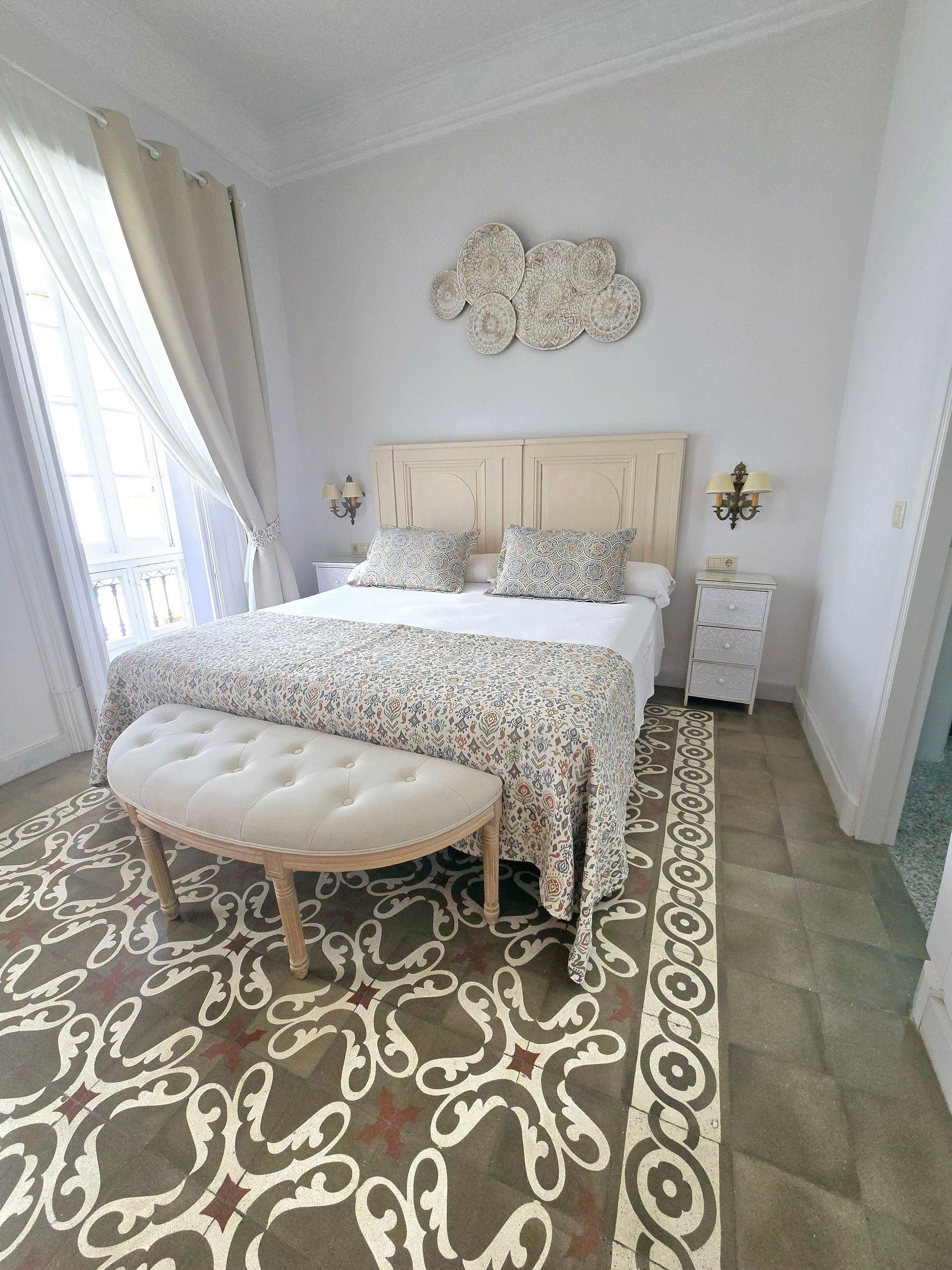 Dormitorio con cama, banco, alfombra estampada, paredes blancas y nube decorativa.