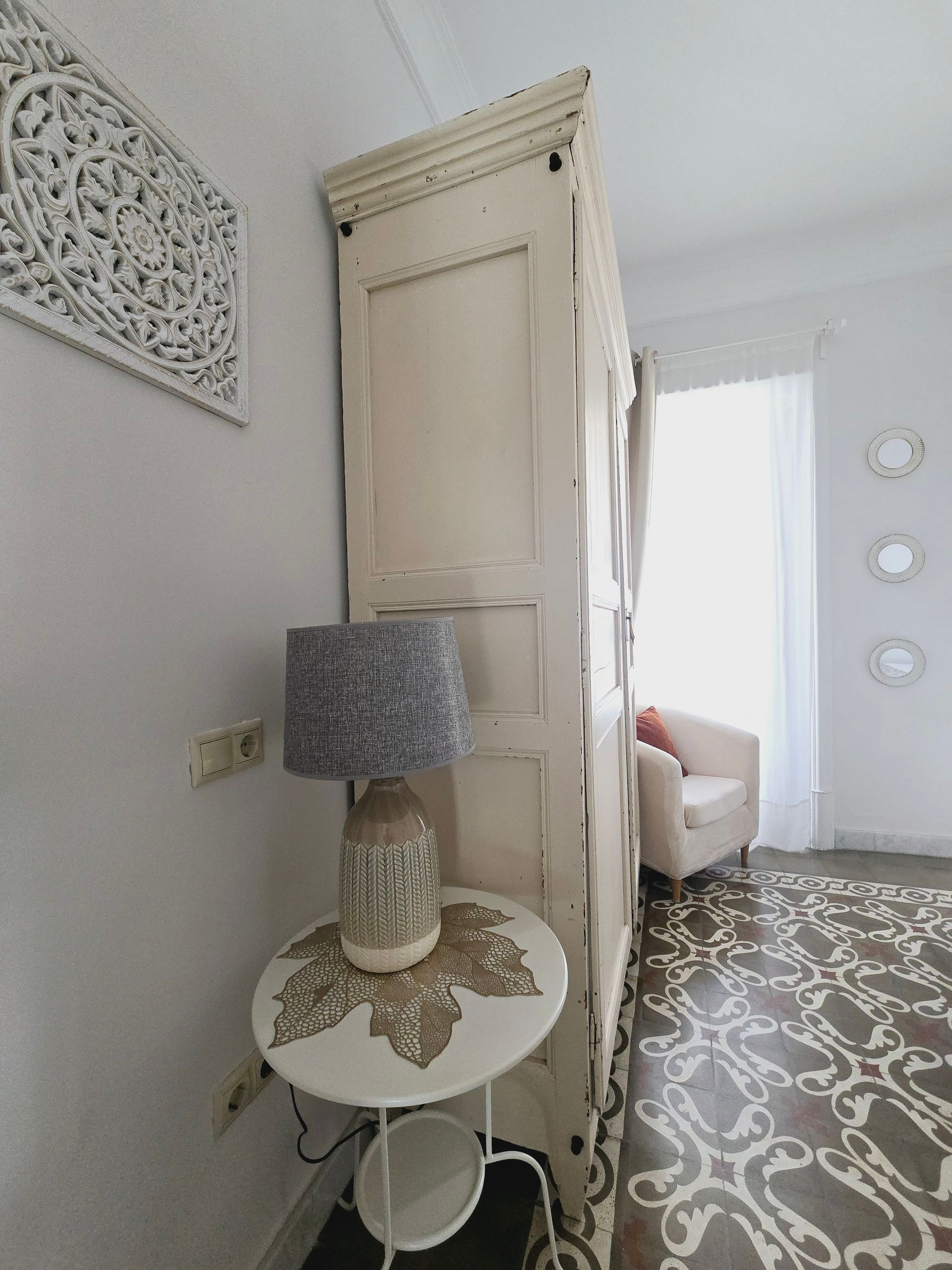 Un armario vintage blanco separa una habitación con suelo estampado. Una lámpara reposa sobre una mesa pequeña.