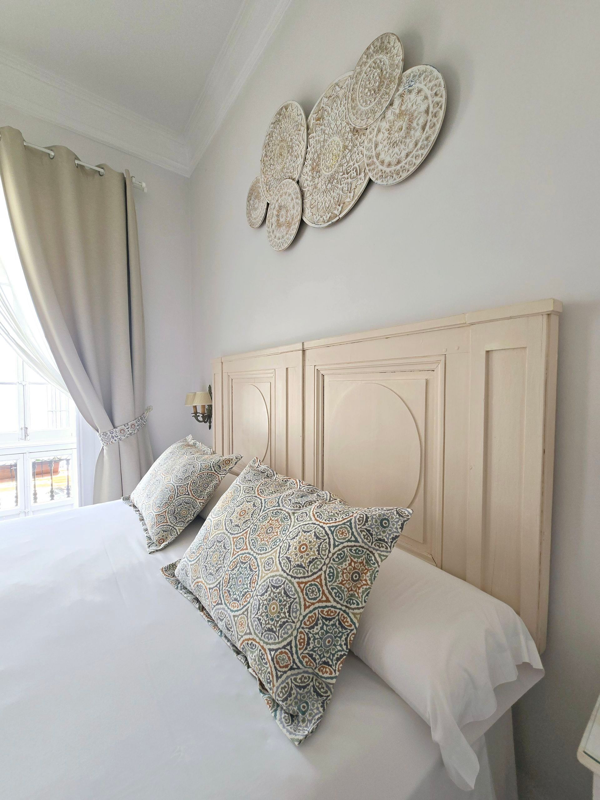 Dormitorio con cabecera de color crema, ropa de cama blanca, almohadas florales y arte de pared en forma de nubes.