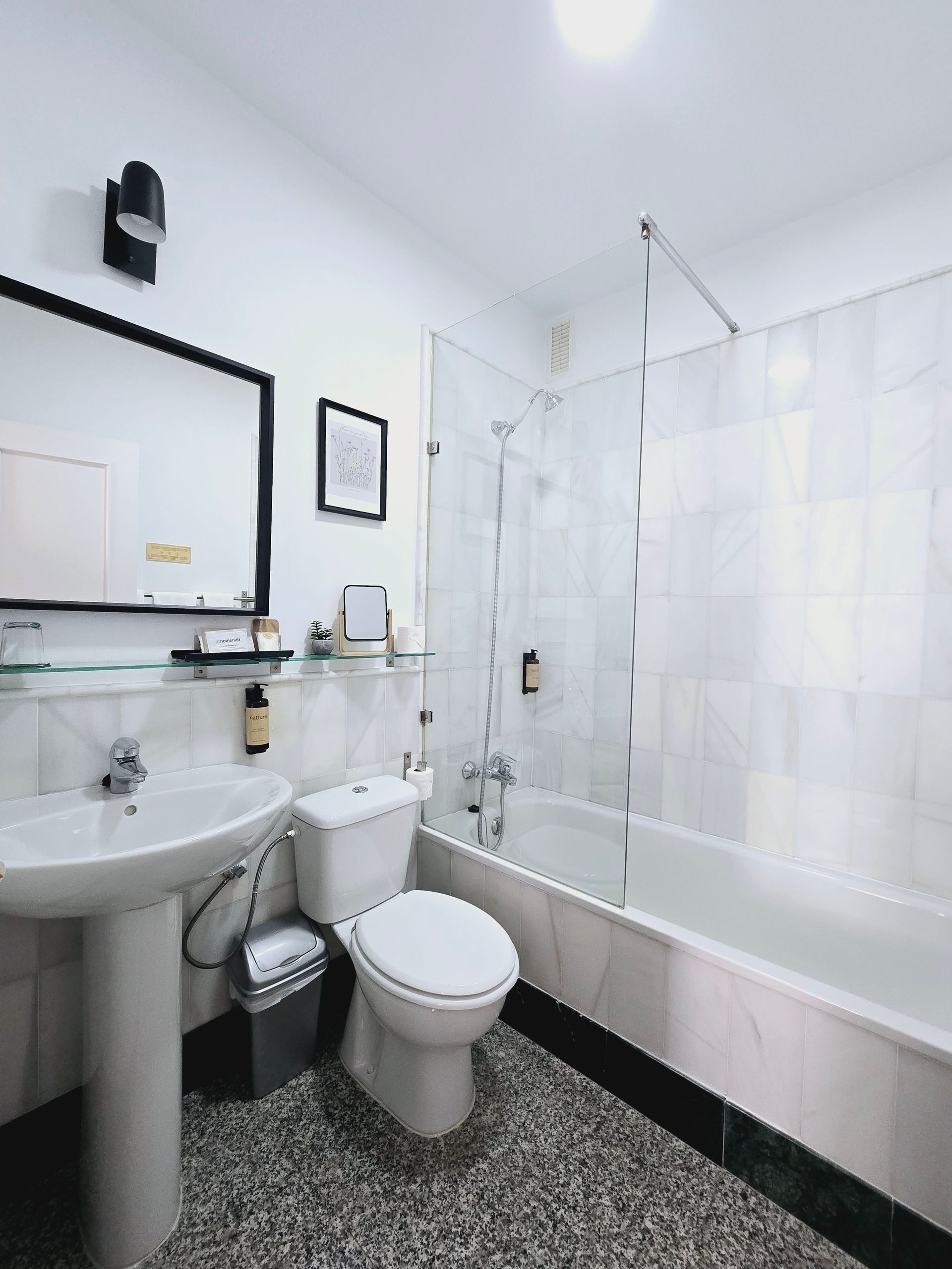 Baño con lavabo, inodoro, bañera y mampara. Paredes blancas, molduras negras y suelo moteado.