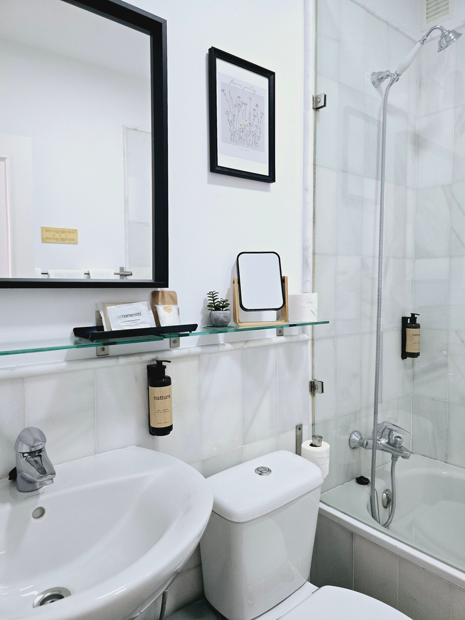 Baño blanco con lavabo, inodoro y ducha. Espejo, obras de arte y artículos de aseo en un estante.