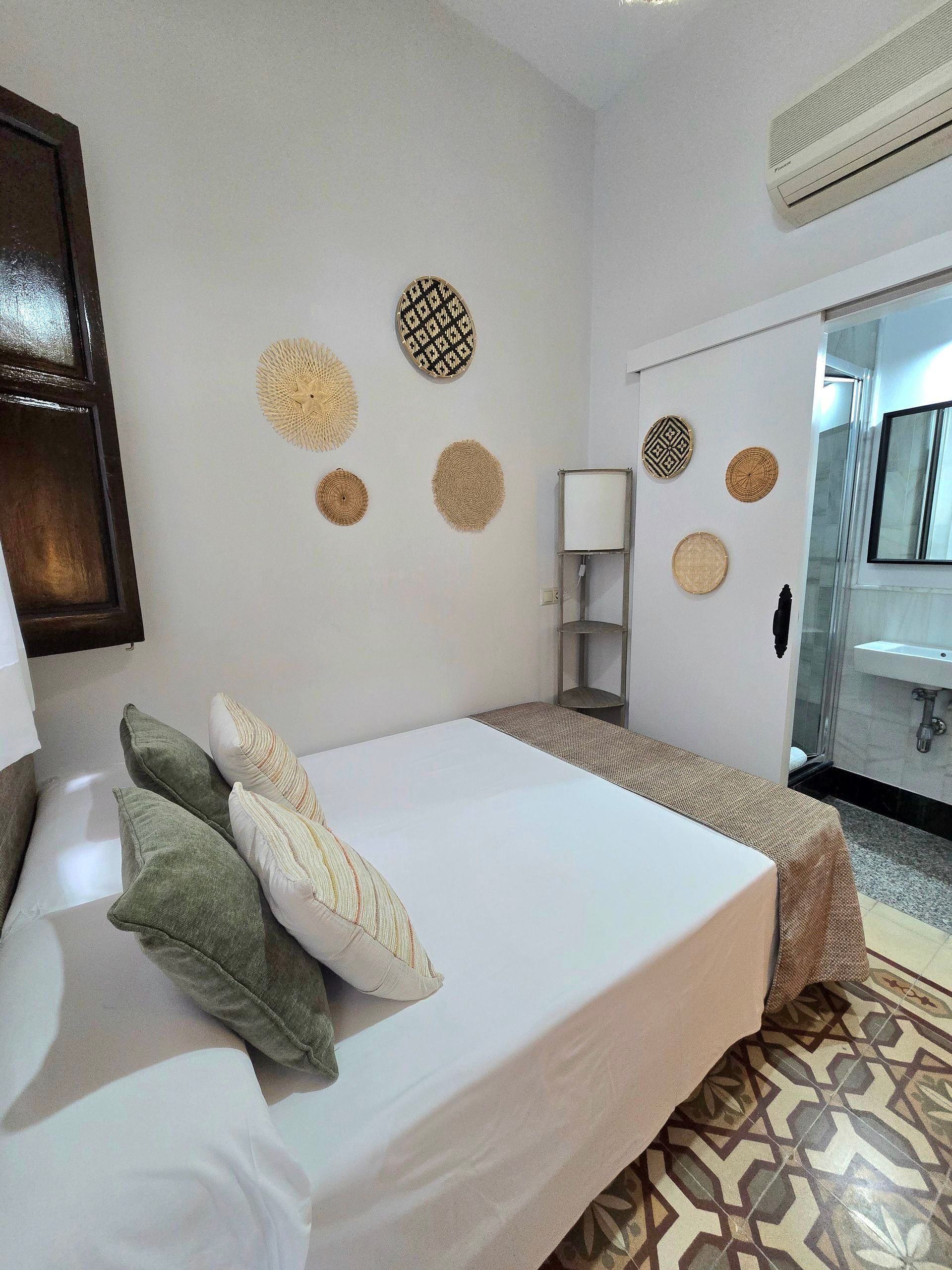 Dormitorio con ropa de cama blanca, cestas decorativas en la pared y vista a un baño con puerta corrediza.