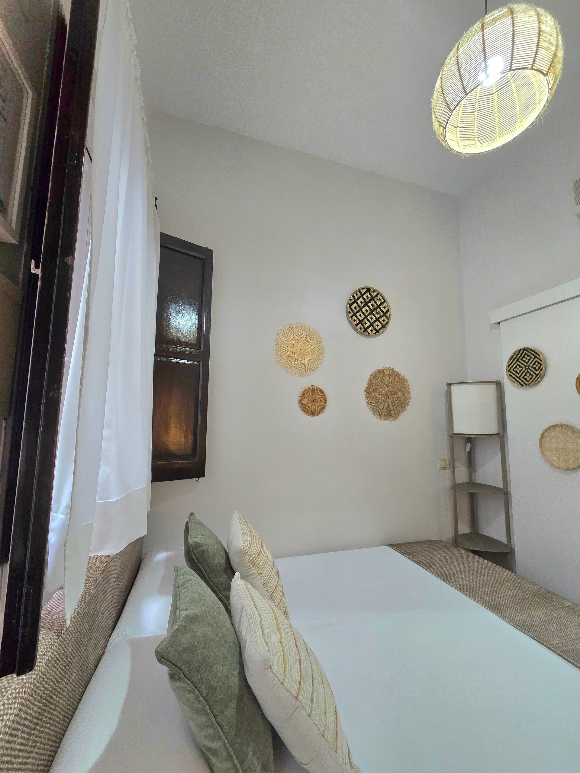 Dormitorio con paredes blancas, cama, ventana, decoración de pared tejida y una lámpara de techo estampada.