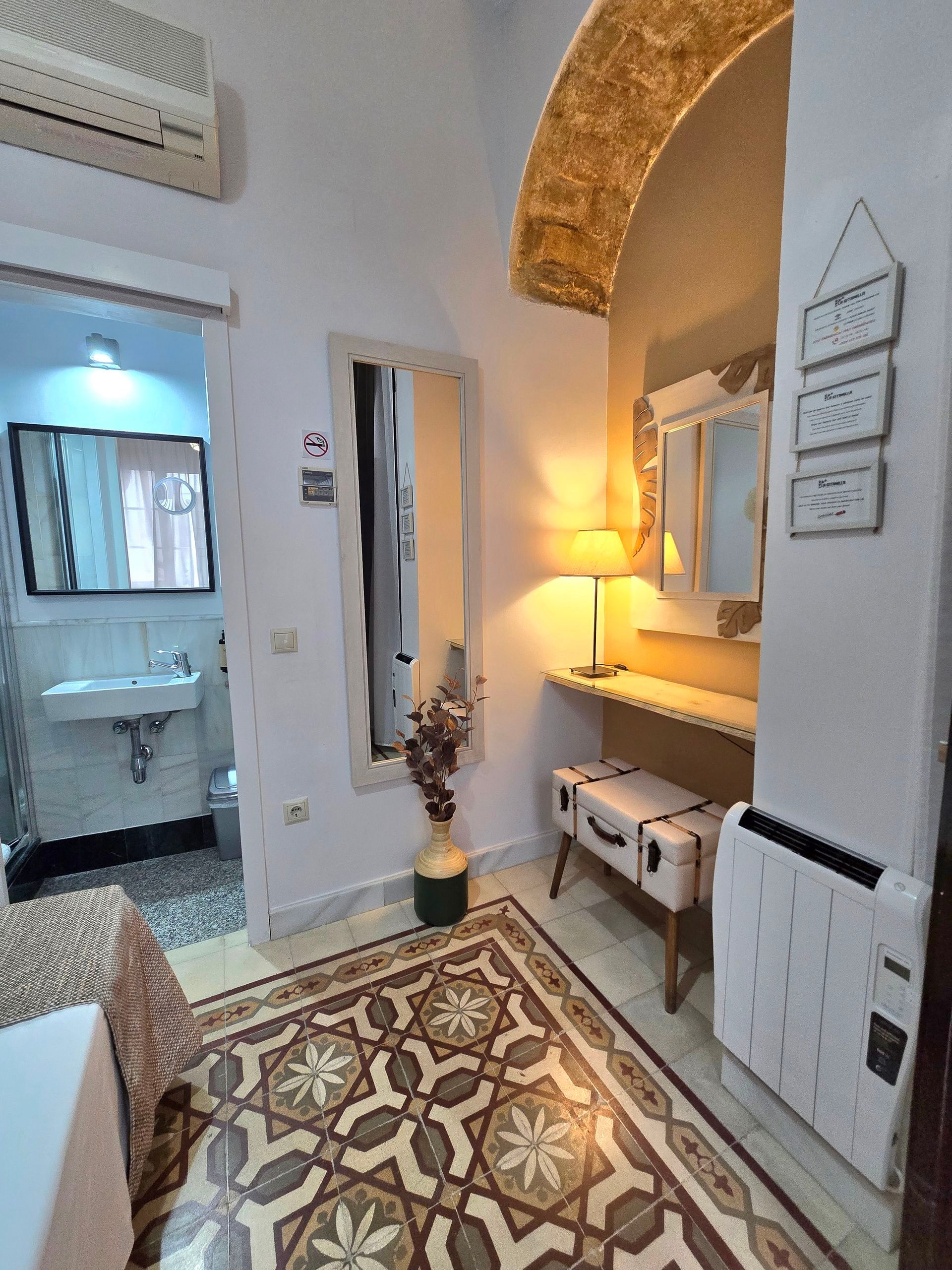 Pasillo con piso de baldosas estampadas, espejo, tocador y puerta de acceso a un baño.