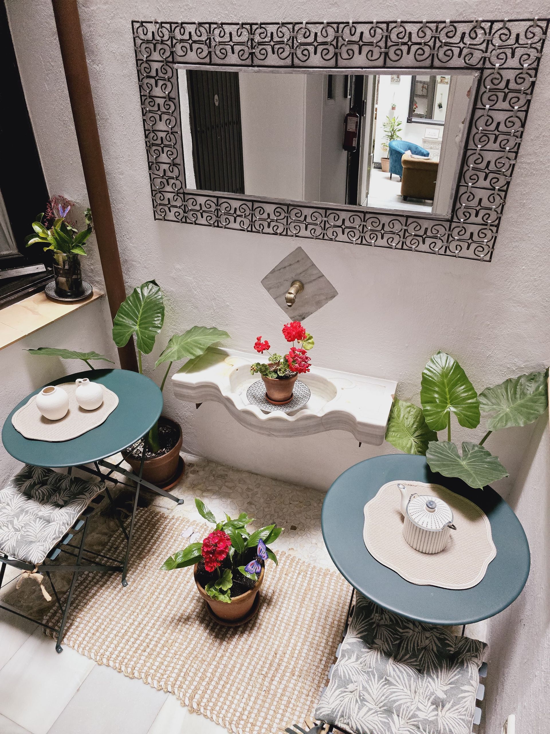 Pequeño patio al aire libre con dos mesas color verde azulado, plantas y un espejo.