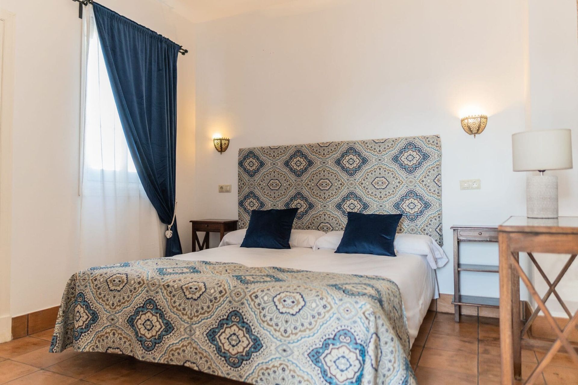 Dormitorio con cabecero, colcha y cortinas con estampados azules y blancos. Paredes beige y muebles de madera.