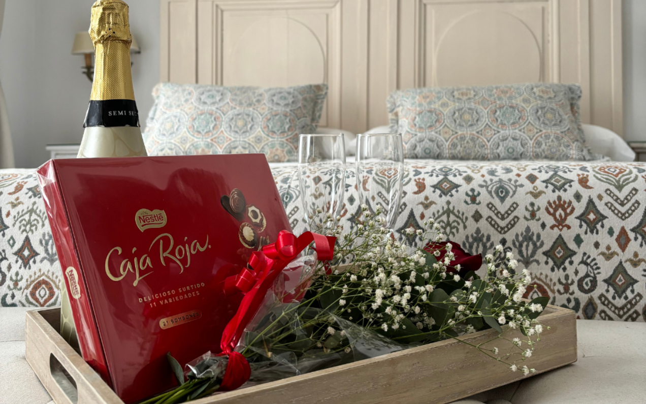 Bandeja con bombones, champán, flores y copas sobre una cama con almohadas estampadas.