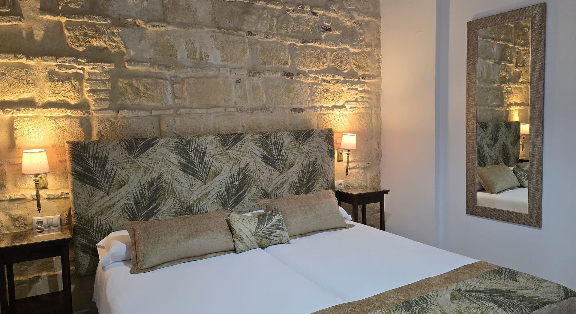 Dormitorio con pared de piedra, cama, lámparas, mesitas de noche y espejo.