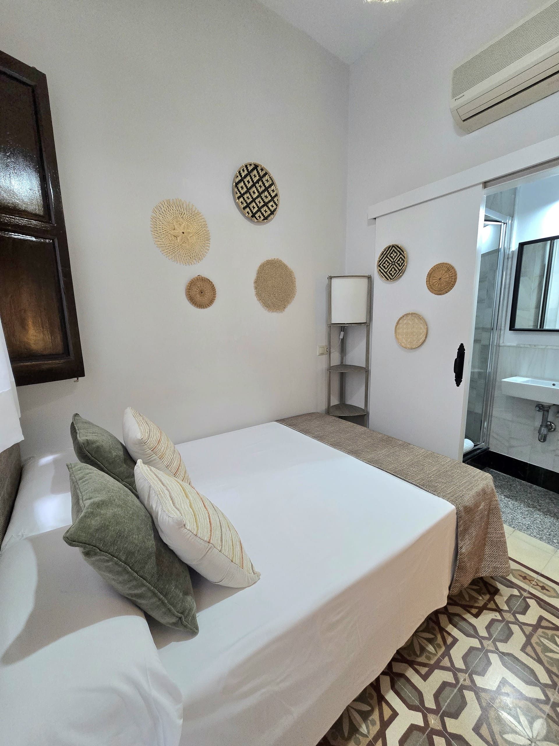 Dormitorio con paredes blancas, cama con almohadas y arte decorativo. Baño visible a través de una puerta corredera.