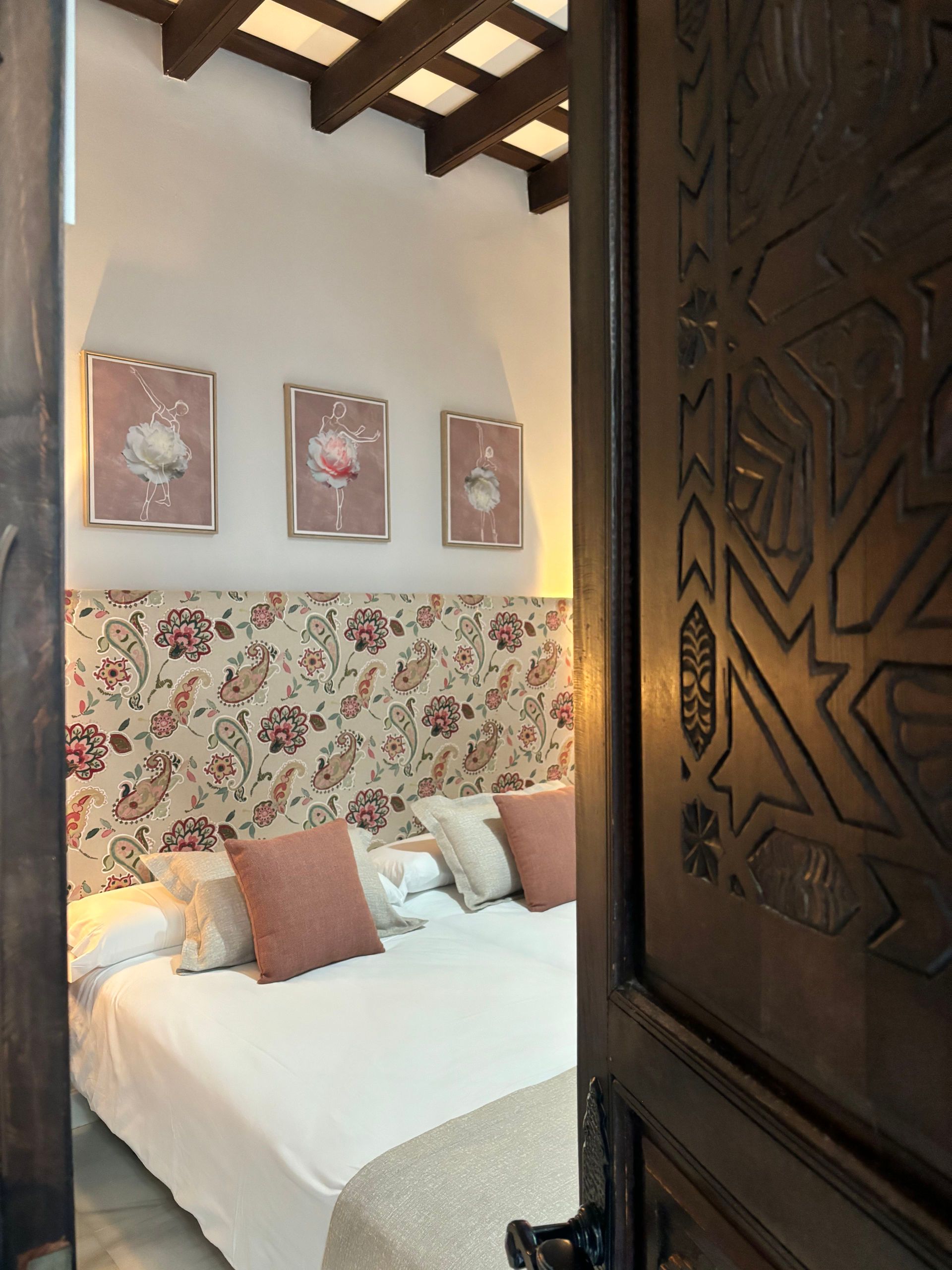 Interior de dormitorio con cabecera floral, arte enmarcado y puerta de madera tallada decorativa.