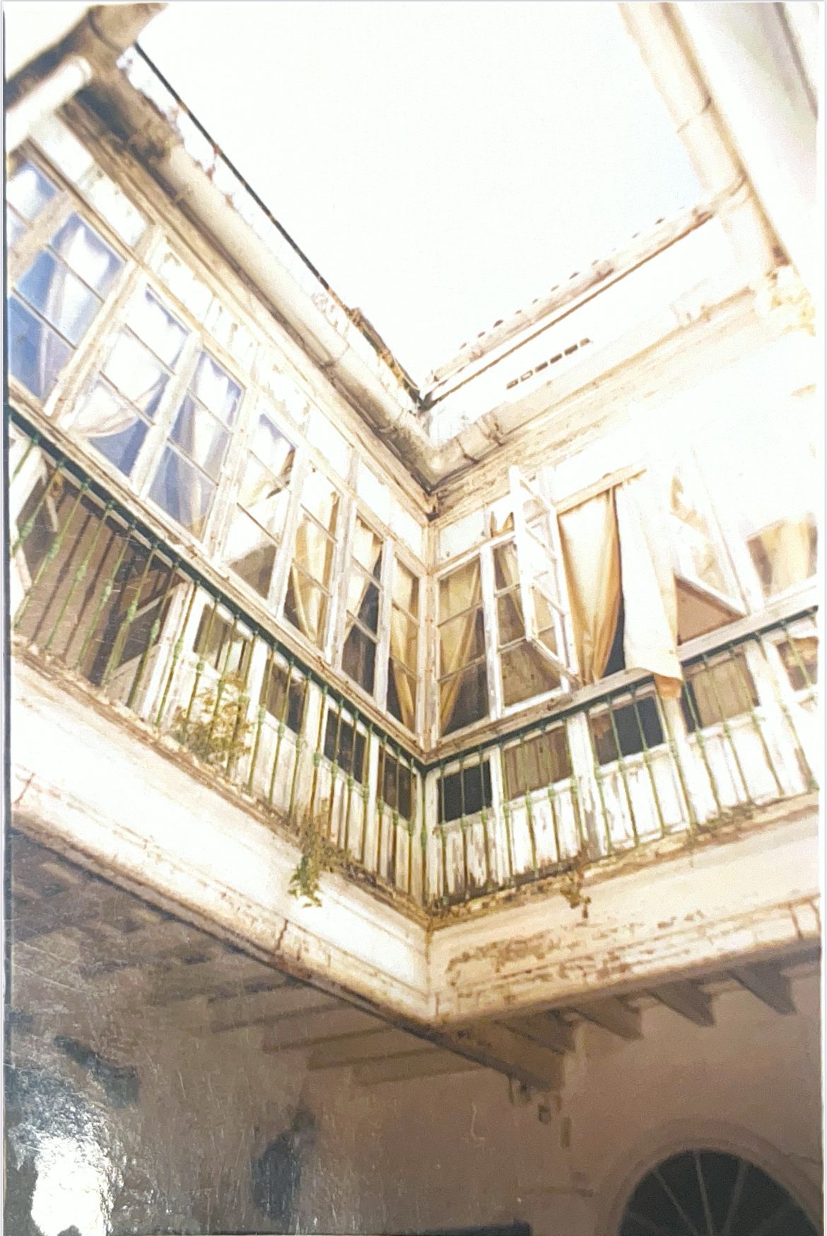 Mirando hacia arriba a un balcón esquinero blanco y desgastado con múltiples ventanas y cortinas.